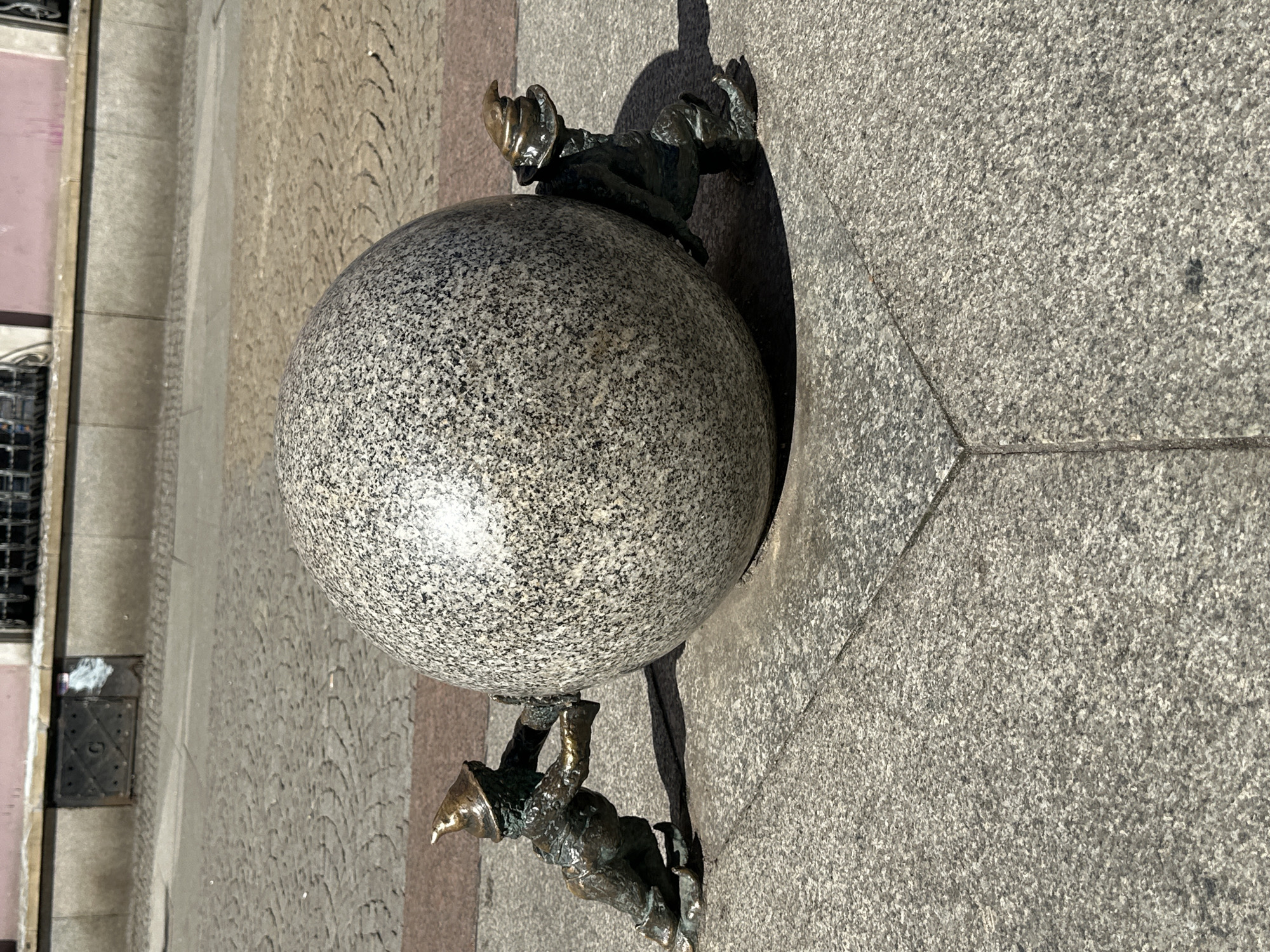 The image shows a granite sphere supported by two bronze dwarf statues, a tourist attraction located in Wrocław, Poland. One dwarf appears to be pushing the sphere from below, while the other is on top, seemingly helping to push it forward. The sphere is sitting on a light gray pavement. The background includes the corner of a building with a window and parts of the paved street. The scene is illuminated by bright sunlight.