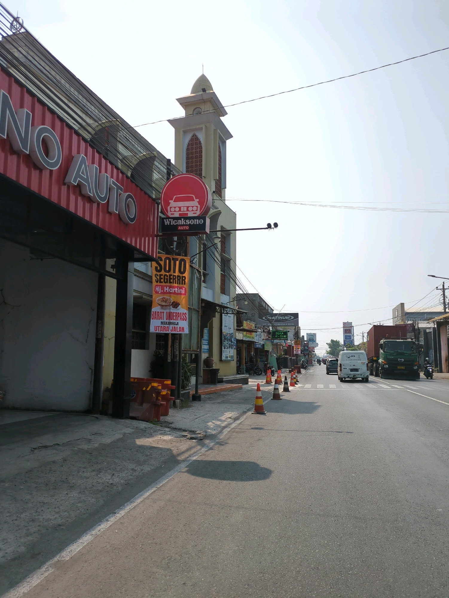 The image depicts a street scene in Pabelan, Indonesia, under a bright, clear sky. On the left, a building with a red facade and large white lettering that reads "NO AUTO" is visible, with a section of what appears to be a car wash or auto service bay. A yellow and white striped tower with a dome-like structure stands behind this building. Several signs are present: a circular red sign with a car silhouette and the text "Wicaksono AUTO" is suspended, and a vertical banner below it advertises "SOTO SEGERRR Hj. Hartini," with further text indicating "BARAT UNDERPASS" and "UTARA JALAN."
The street is paved with asphalt and has white lane markings. Several orange and white traffic cones are placed along the sidewalk and on the road. Traffic is moving in the distance, including a white van and a large green truck. The right side of the street features a row of buildings, some with signs and awnings. The environment suggests a commercial area with businesses and ongoing traffic.