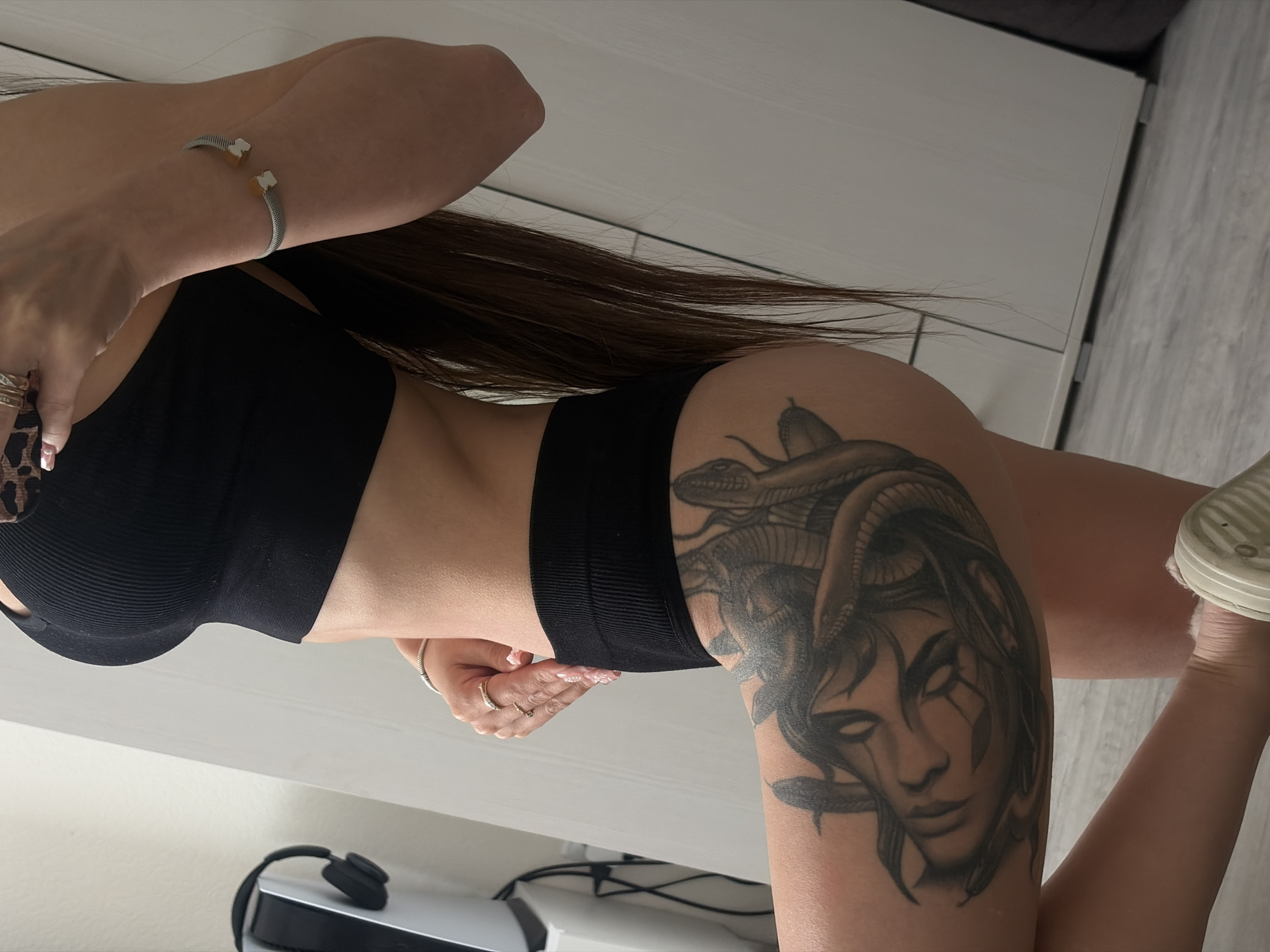 A person, appearing to be female, is depicted from the mid-torso to the knees, kneeling or squatting indoors. She wears a black ribbed athletic crop top and matching high-waisted briefs. Her long, dark brown hair cascades down her back. A silver-toned mesh bracelet with two light-colored bear-shaped charms adorns her left forearm, and multiple rings are visible on the fingers of her right hand, which is resting on her hip. Her fingernails are manicured with a light polish. A large, detailed grayscale tattoo of a Medusa-like face with snakes for hair covers a significant portion of her upper left thigh. Her left foot, wearing a light-colored, possibly off-white, casual shoe, is visible in the lower left corner. The background consists of a plain light-colored wall, possibly a closet or wardrobe with vertical panel lines. To the left, a white and silver electronic device, resembling a gaming console (such as a PlayStation 5), has black over-ear headphones resting on top of it. The flooring is light-colored wood or laminate. The lighting suggests an indoor environment. The indoor setting is situated in the city of Unknown, Russia.