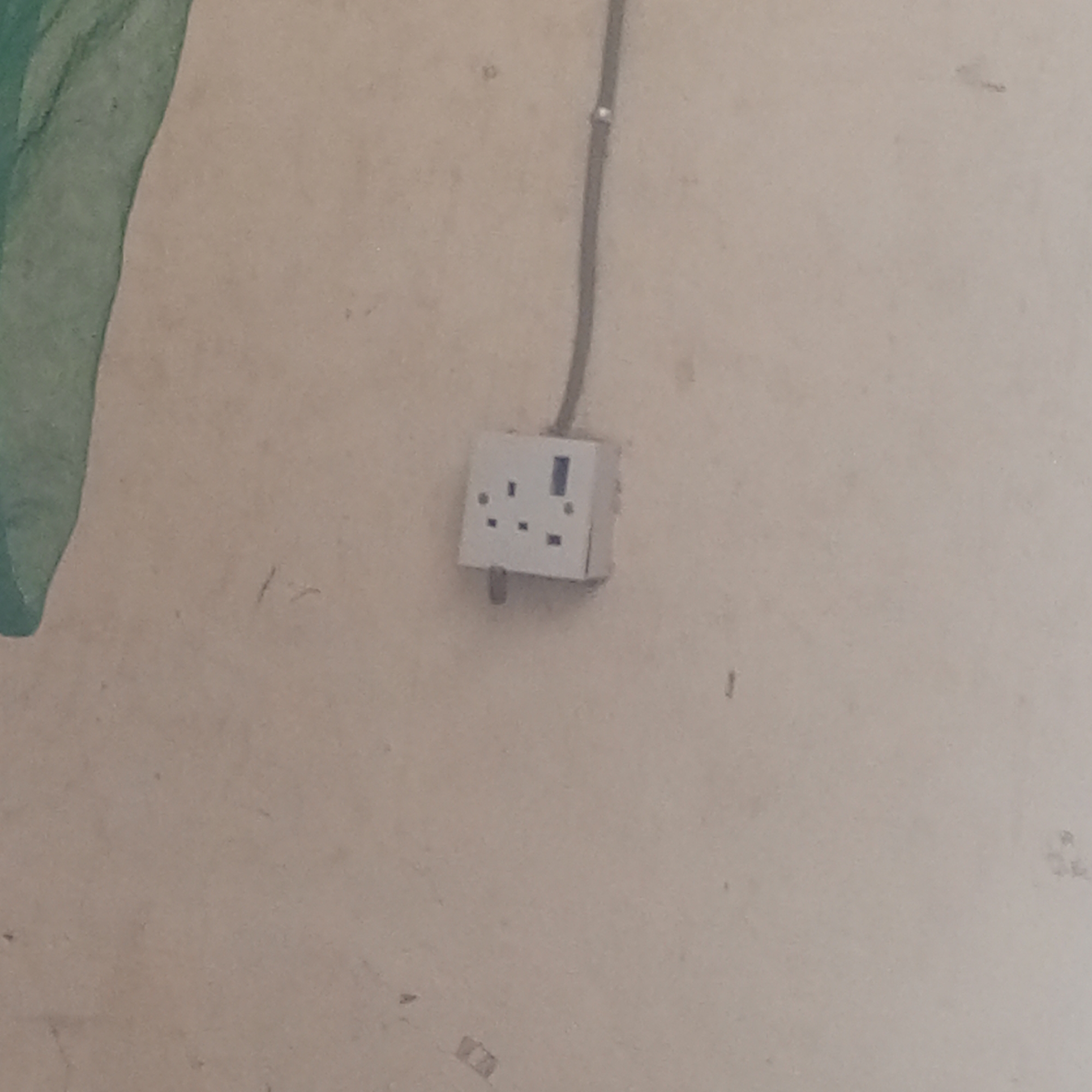 The image shows a close-up of a white electrical outlet mounted on a light-colored wall. The outlet is a universal type with three rectangular openings and one round opening, suggesting it is designed to accept various plug types. A gray electrical cable runs from the top of the outlet upwards, disappearing out of frame. A portion of a green, possibly fabric or netting material, is visible on the left side of the image, partially obscuring the wall. The wall itself appears to be textured and shows some signs of age or wear, with faint marks and smudges. The lighting is neutral, indicating it's likely indoors during daylight or under artificial light. There are no people or other discernible objects in the immediate vicinity of the outlet. The overall scene is unremarkable, focusing solely on the electrical fixture and its immediate surroundings. The location context provided is Zinna, Nigeria.