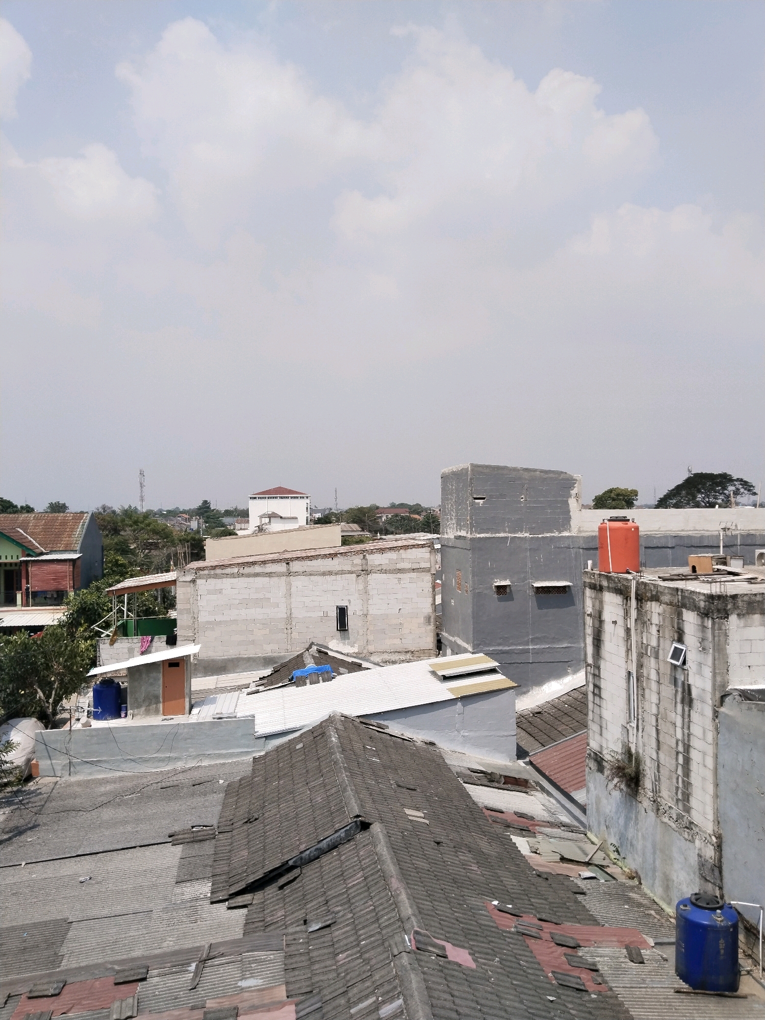 The image captures a rooftop view of a densely built urban area in Jalenjaya, Indonesia. In the foreground, a heavily tiled roof with sections of rust-colored roofing material shows signs of wear and tear. To the left, a lighter colored corrugated roof is visible. Beyond this, multiple buildings are seen, constructed from various materials including grey concrete blocks and painted concrete.
Prominently featured is a tall, dark grey rectangular building with several small, rectangular openings and a larger rectangular window. To its right, an orange water tank is situated on top of a lower, white-roofed structure. Further to the right, another building constructed with white concrete blocks has a weathered appearance and a small, open window.
In the midground, a building with a white facade and a red-tiled roof stands out. Other structures, some appearing to be under construction or renovation with exposed concrete blocks, are interspersed with trees. A blue water tank is visible on a lower roof to the left. In the distance, a telecommunications tower can be seen rising above the treeline. The sky is mostly overcast with patches of white clouds. The scene depicts a typical residential and commercial rooftop landscape in a developed urban environment.