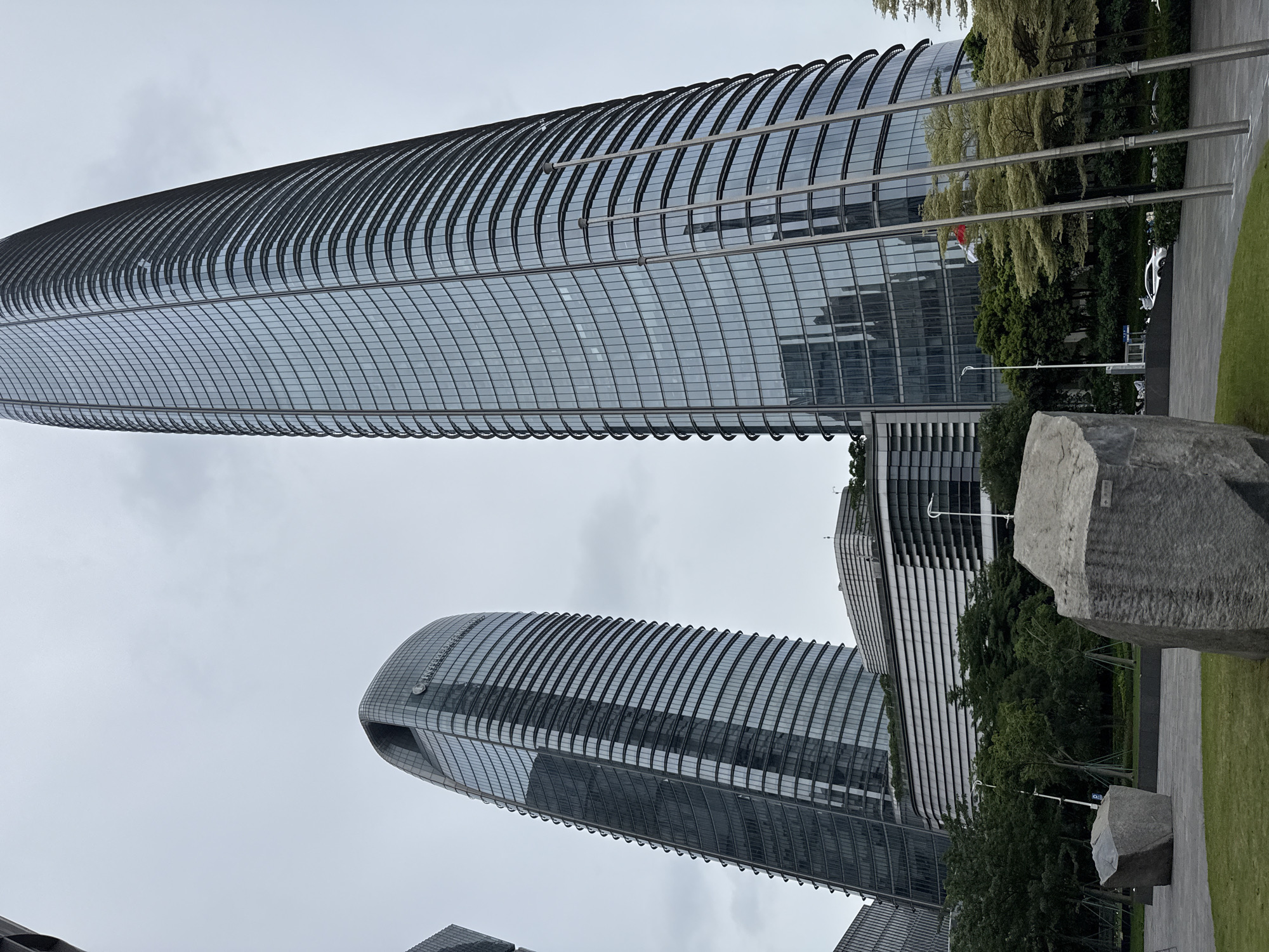 The image displays the exterior of two modern skyscrapers in Haizhu District, China. The taller, rightmost building is a sleek, curved glass structure with numerous horizontal lines created by its facade. To its left is a shorter, similarly curved skyscraper with a distinctive arched top, also clad in reflective glass. In the foreground, two large, rough-hewn gray rocks are positioned on a manicured lawn and paved area. Behind the rocks, a low-rise building with a distinctive layered facade is visible, along with a cluster of lush green trees. The sky is overcast and appears a pale gray. Several flagpole-like structures are also present in the midground. A white car is partially visible in the background, near the low-rise building. The scene suggests an urban environment with a focus on modern architecture and public space.