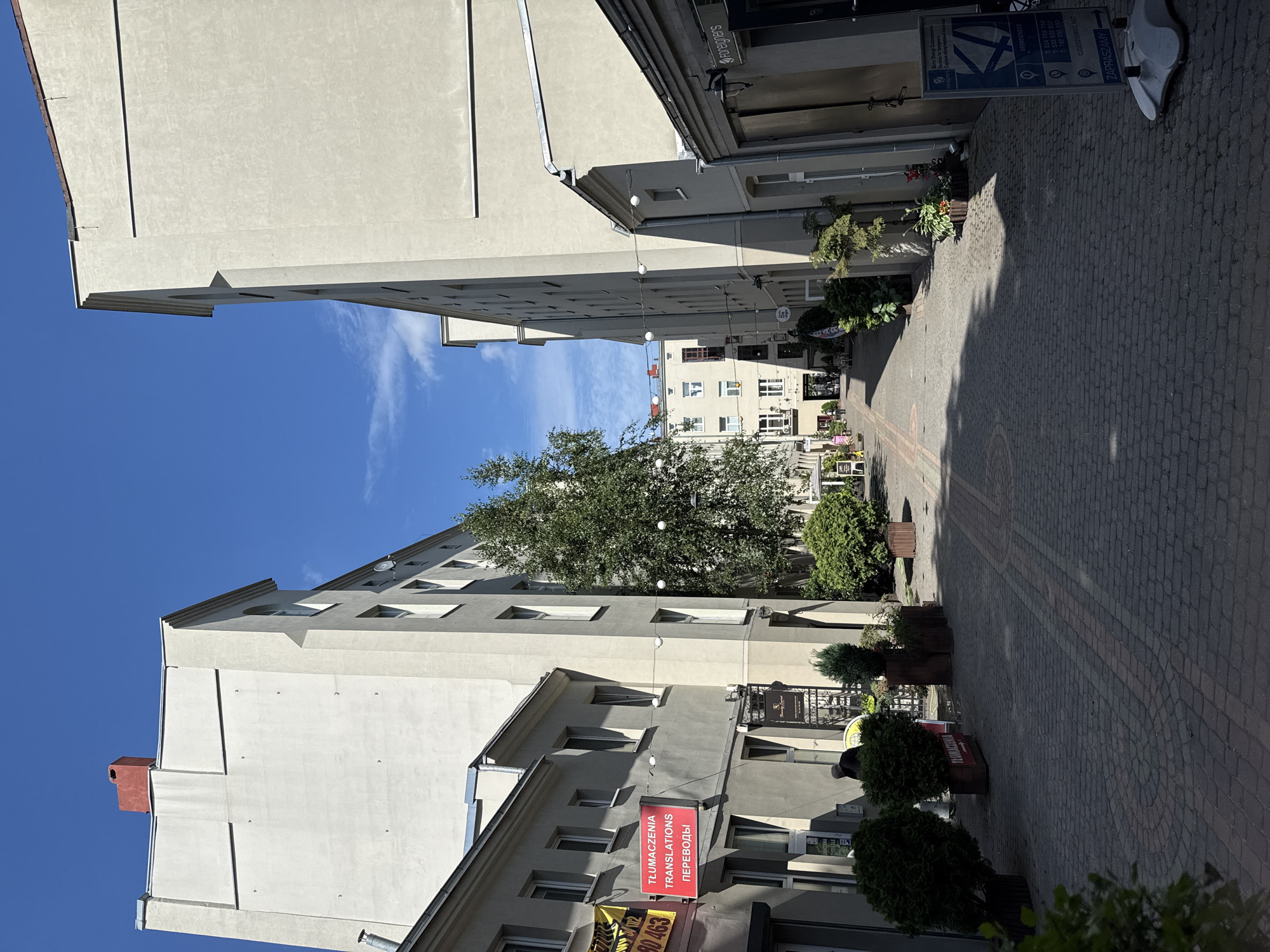 The image shows a narrow street between two buildings in Łódź, Poland, likely in the city center. The street is paved with cobblestones and lined with trees and shrubs. The buildings are multi-story and appear to be a mix of residential and commercial spaces. On the left side of the street, a red sign with white lettering reads "TŁUMACZENIA TRANSLATIONS ПЕРЕВОДЫ," indicating translation services. There is also a yellow sign with black text. A person, partially visible, is standing near the entrance of one of the buildings. On the right side of the street, a blue sign with white text and graphics provides information about services for foreigners. The sky is blue with some scattered clouds, suggesting fair weather. The overall impression is one of a quiet, possibly historic, urban environment.