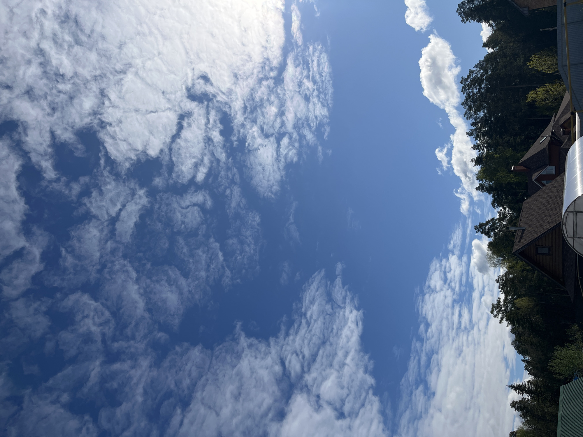 The image captures a view of a partly cloudy sky over the city of Chaikovsky, Russia. The sky is predominantly blue with scattered formations of white, fluffy clouds. Along the right edge of the image, a portion of a building's roof is visible, along with some trees. The overall scene suggests a bright, sunny day in a residential or suburban area.