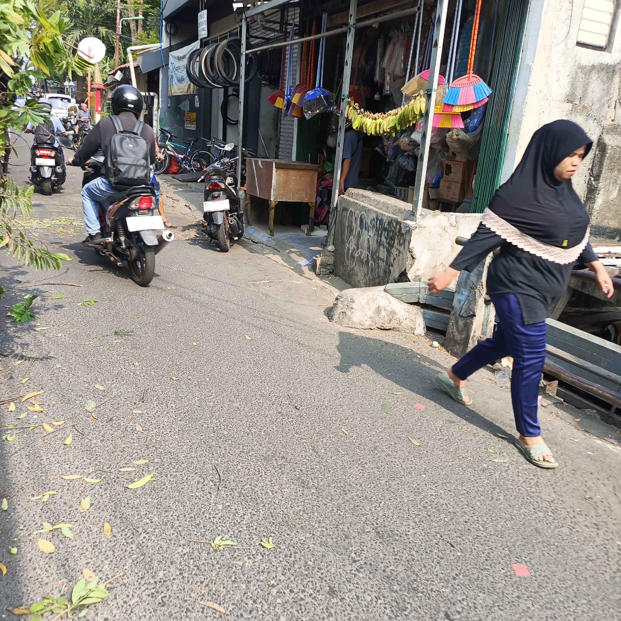 The image depicts a street scene in South Jakarta, Indonesia, under bright daylight. In the foreground, a woman wearing a black hijab and dark blue pants is walking away from the camera, her body angled towards the right. She is wearing light green sandals. Behind her and to the left, a motorcycle with a rider wearing a helmet and backpack is moving away from the camera on the asphalt road. Further down the street, another motorcycle is visible, also moving away. The street is lined with shops, one of which has an open front displaying various items, including brooms with colorful handles and bicycle tires mounted on the wall. The pavement is littered with fallen leaves and twigs. A concrete structure with graffiti is present on the right side of the road, partially obscuring a metal railing. The environment suggests a busy urban area with commercial activity.
