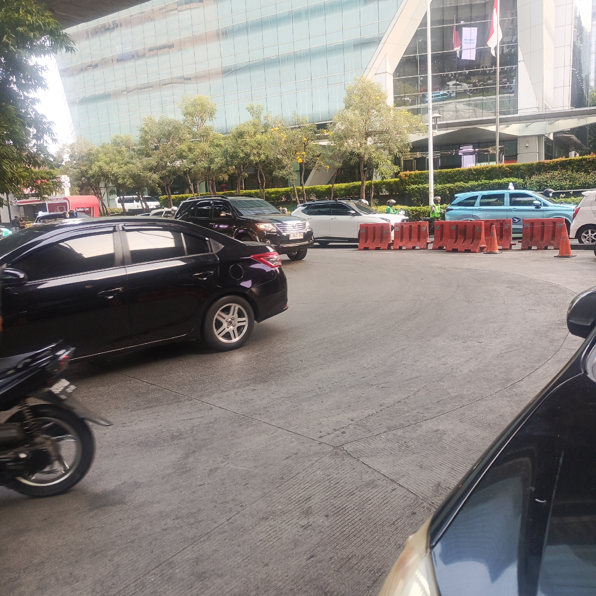 A traffic scene unfolds on a paved road in South Jakarta, Indonesia, with a large, modern glass building dominating the background. The building's facade reflects the sky and surrounding trees, creating a dynamic visual effect. In the foreground, a black sedan with tinted windows is parked, its rear wheel clearly visible. To the right, a dark-colored car's hood and headlight are partially in frame.
The road is lined with orange traffic barriers, some of which are upright cones. Several vehicles are present: a dark SUV is stopped behind the sedan, and a white SUV is further ahead. A light blue van, possibly a taxi, is also present. Two individuals wearing green vests and helmets, likely traffic personnel or delivery riders, are positioned near the blue van. A motorcycle is visible to the left, its rear wheel blurred by motion. Trees and manicured bushes line the area in front of the building, adding a touch of greenery to the urban landscape. The overall scene suggests a busy street with ongoing traffic flow.