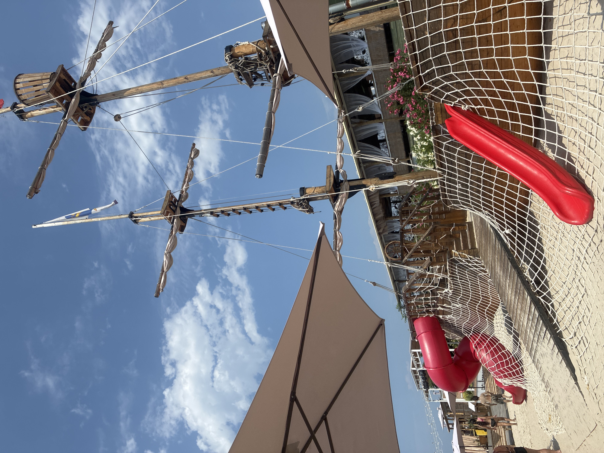 A sunny day at a beachfront playground in Sirius, Russia, features a wooden pirate ship structure on a sandy area. Two red slides, one large curved one and a smaller straight one, are integrated into the ship's design, connected by a wooden ramp and white netting. In the background, a covered seating area with white curtains and pink flowers is visible. Several people are present, some walking on the sand near the playground. The sky is blue with scattered white clouds.