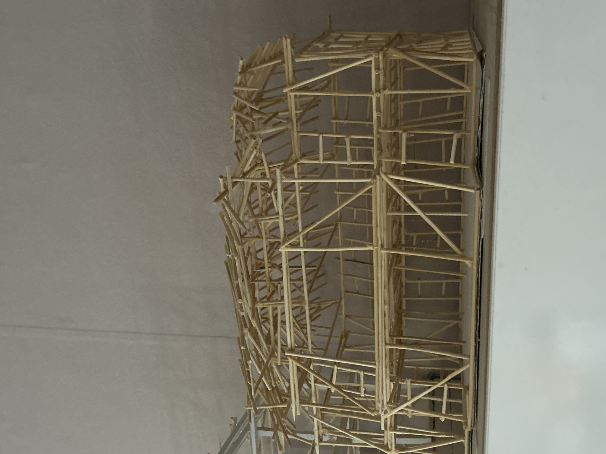 The image depicts a model of a building or structure constructed from thin wooden sticks, possibly matchsticks or toothpicks. The model has a somewhat chaotic appearance, with many of the sticks arranged in an irregular or haphazard fashion. The model appears to be placed in the corner of a room, with a plain wall in the background on the left and a white or light-colored surface on the right. The lighting is somewhat dim and even, making it difficult to discern fine details. The image does not provide any information about the location of the model in the city of Sofia, Bulgaria.
