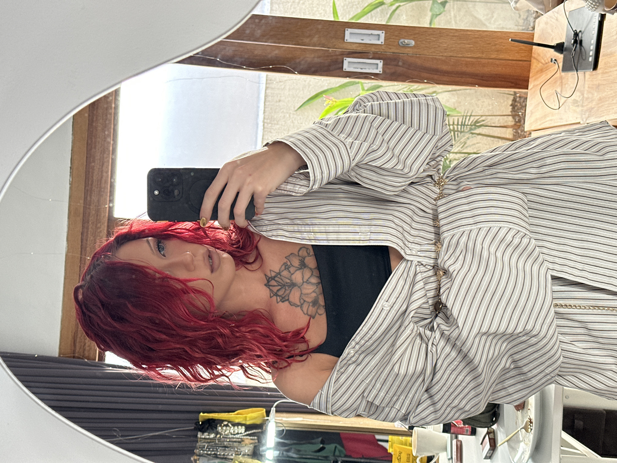 The image displays a mirror selfie of a woman with vibrant red, wavy hair. She is captured from the chest up, with her reflection showing her holding a black smartphone in her right hand to take the picture. Her fingernails are painted gold. She has light-colored eyes and a nose piercing, with a neutral expression on her face. She is wearing a black top or bra underneath a loosely draped, unbuttoned striped shirt, which features thin vertical stripes in light brown/beige and white. A prominent floral and geometric tattoo is visible on her left upper chest and shoulder. A subtle gold chain accessory is also noticeable.
The reflection in the irregularly shaped mirror reveals an indoor setting bathed in bright natural light, indicating daytime. The background includes a wooden surface, possibly a desk or a set of drawers, with two small white label holders. Green foliage with a small pink flower suggests the presence of plants, either indoors or visible through a window. A black electronic device with an antenna and wires sits on a wooden surface. Various objects, including what appears to be stacked jewelry or bracelets and a yellow container with faint text, are reflected on a lower surface. The scene is a personal snapshot taken in North Kuta, Indonesia.
