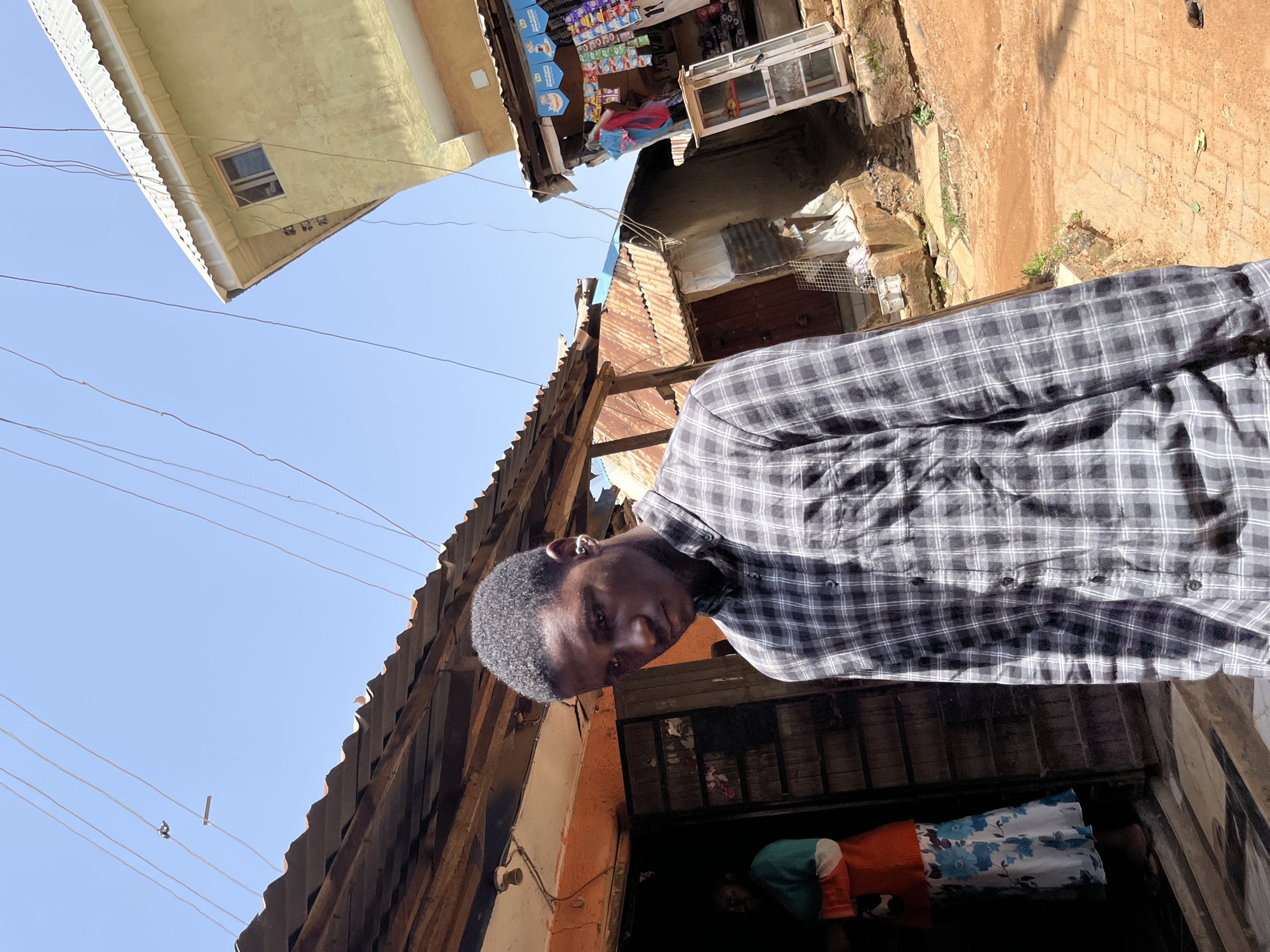 The media file shows a young Black male in the foreground, wearing a gray and black checkered button-down shirt. He is looking down and slightly to his left, with an earring visible in his left ear. The background depicts an outdoor scene with several buildings and structures that appear to be in a market or street setting. One building to the left has a yellow facade and a white corrugated roof. In the center, there are stalls with goods displayed, including bags of chips and other packaged items. Further in the background, there are corrugated iron roofs and what looks like a brick-paved area.
The image is taken during the daytime, as indicated by the bright sunlight and clear blue sky. The overall impression is that of a bustling urban environment, possibly in Jos, Nigeria, given the context. The lighting suggests it could be late morning or early afternoon. The man's expression is neutral, and the scene appears to be a candid moment rather than a staged event. There is a second person visible in the lower right portion of the image, wearing an orange shirt and a patterned skirt or dress. The structures suggest a mix of older and newer construction, with some materials appearing weathered. No visible text is clearly legible in the image.