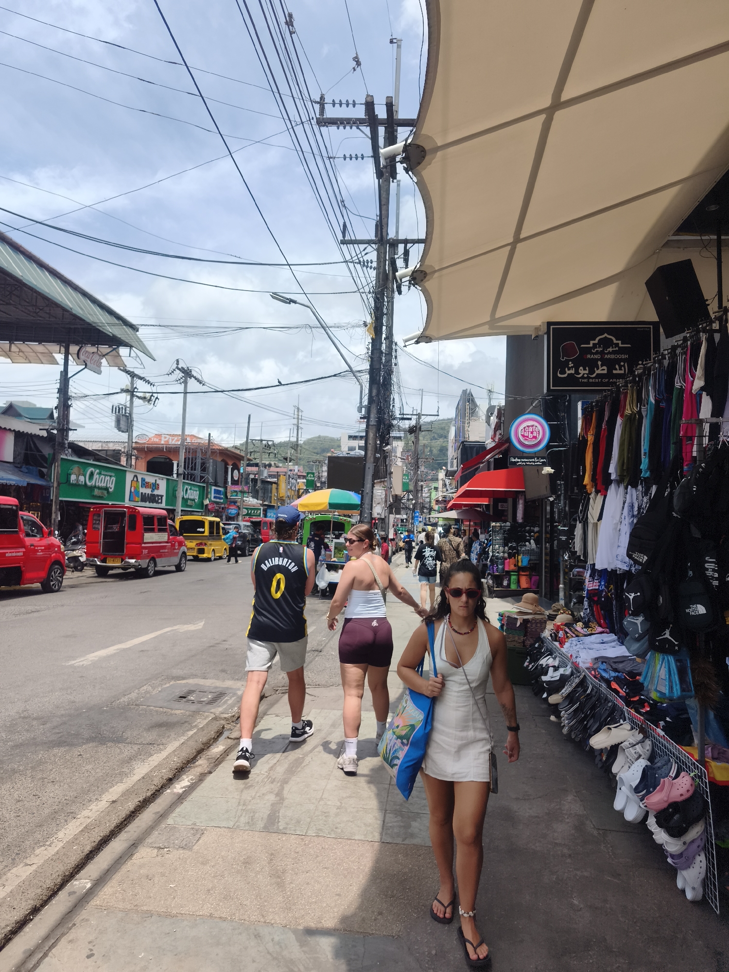 In Patong, Thailand, a street scene unfolds under a cloudy sky, with a road and sidewalk occupied by pedestrians and vehicles. On the sidewalk to the right, a woman in a white dress and sandals walks towards the viewer, carrying a blue patterned tote bag. Behind her, two people walk away from the camera, one in a black and yellow basketball jersey and shorts, the other in athletic shorts and a tank top. Shops line the right side of the street, displaying clothing and accessories. Red and yellow vehicles are visible on the road to the left. Utility poles and wires crisscross overhead, indicating a densely populated urban environment. The overall atmosphere is one of a bustling commercial street.