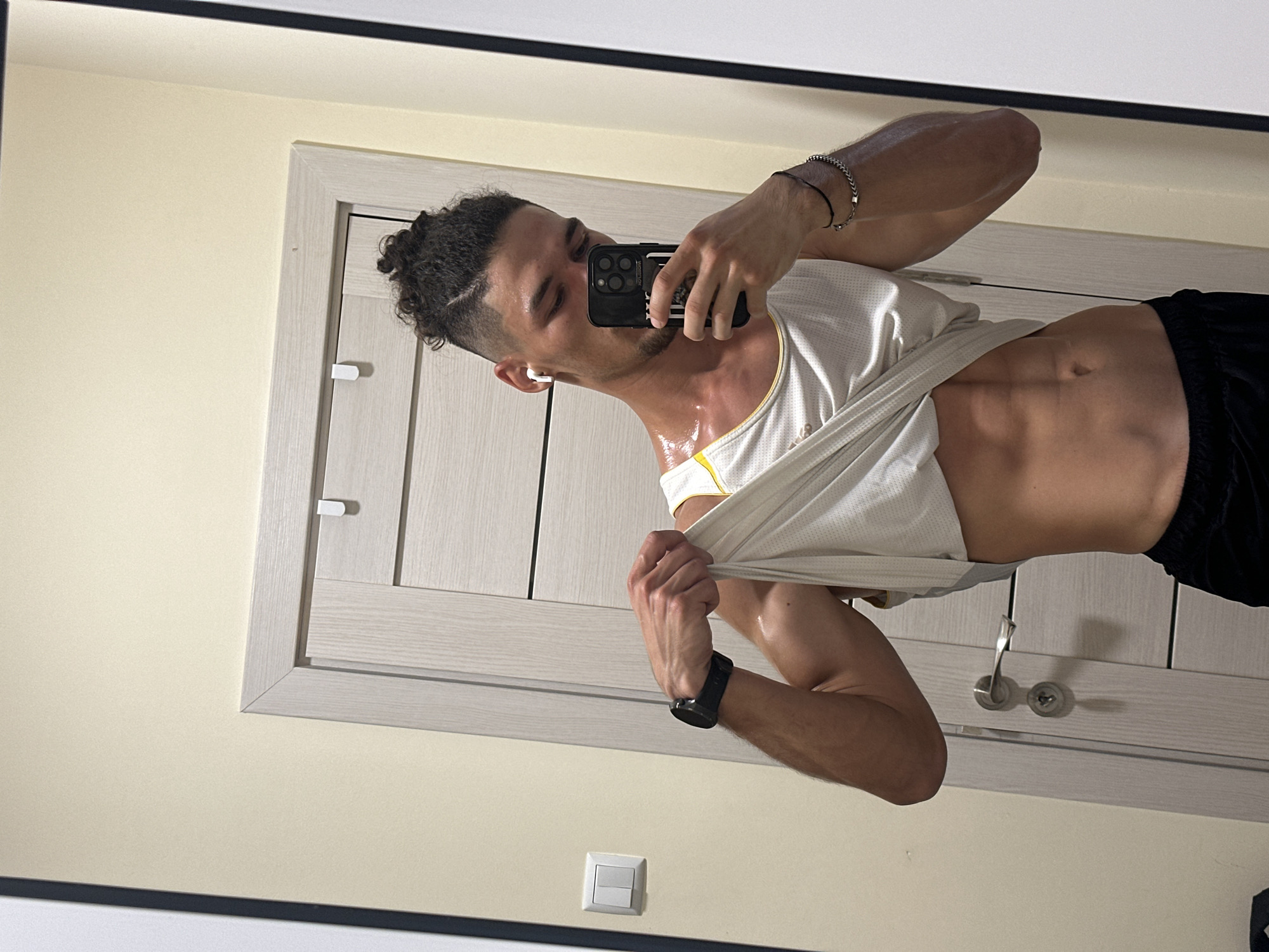A young man with curly hair in a bun, wearing a white tank top and black shorts, takes a mirror selfie. He is showing off his abs and has sweat on his skin. He is wearing white earbuds and a smartwatch. He has a bracelet on his left wrist. The background appears to be a room with a light-colored wall, a light wood door with a silver handle, and a light switch.