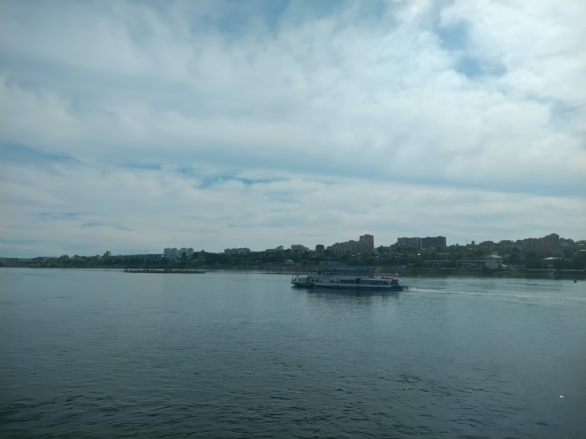 The image depicts a wide, calm river with a white passenger boat with a blue stripe on its side sailing across it. The boat is mid-frame, moving from left to right. In the background, a city skyline with numerous multi-story buildings is visible along the riverbank, suggesting an urban environment. The sky above is partly cloudy with a mix of white and grey clouds against a pale blue backdrop. The overall scene is serene and suggests a pleasant day for a boat ride in a city. This scene is likely of the Angara River in Irkutsk, Russia, which is known for its wide expanse and the city's development along its banks.