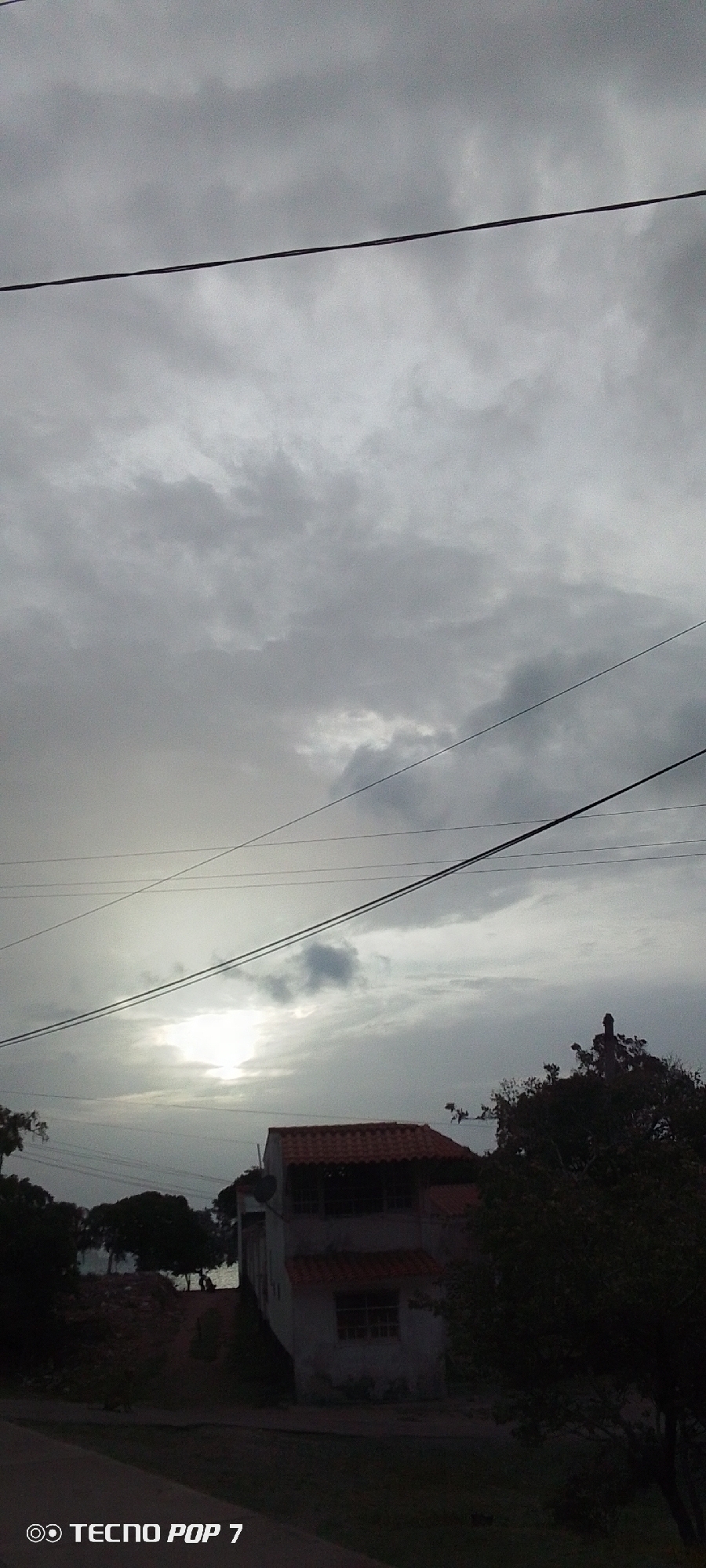 The photograph, seemingly taken in Pedro González, Venezuela, depicts a cloudy, overcast day. A two-story building with a red tile roof is visible in the mid-ground, partially obscured by trees. Overhead, multiple power lines stretch across the frame against the backdrop of a grey, cloud-filled sky, with a diffused light source suggesting the sun trying to break through the clouds. The scene is dimly lit. The 'TECNO POP 7' watermark suggests the image was captured using that model phone.