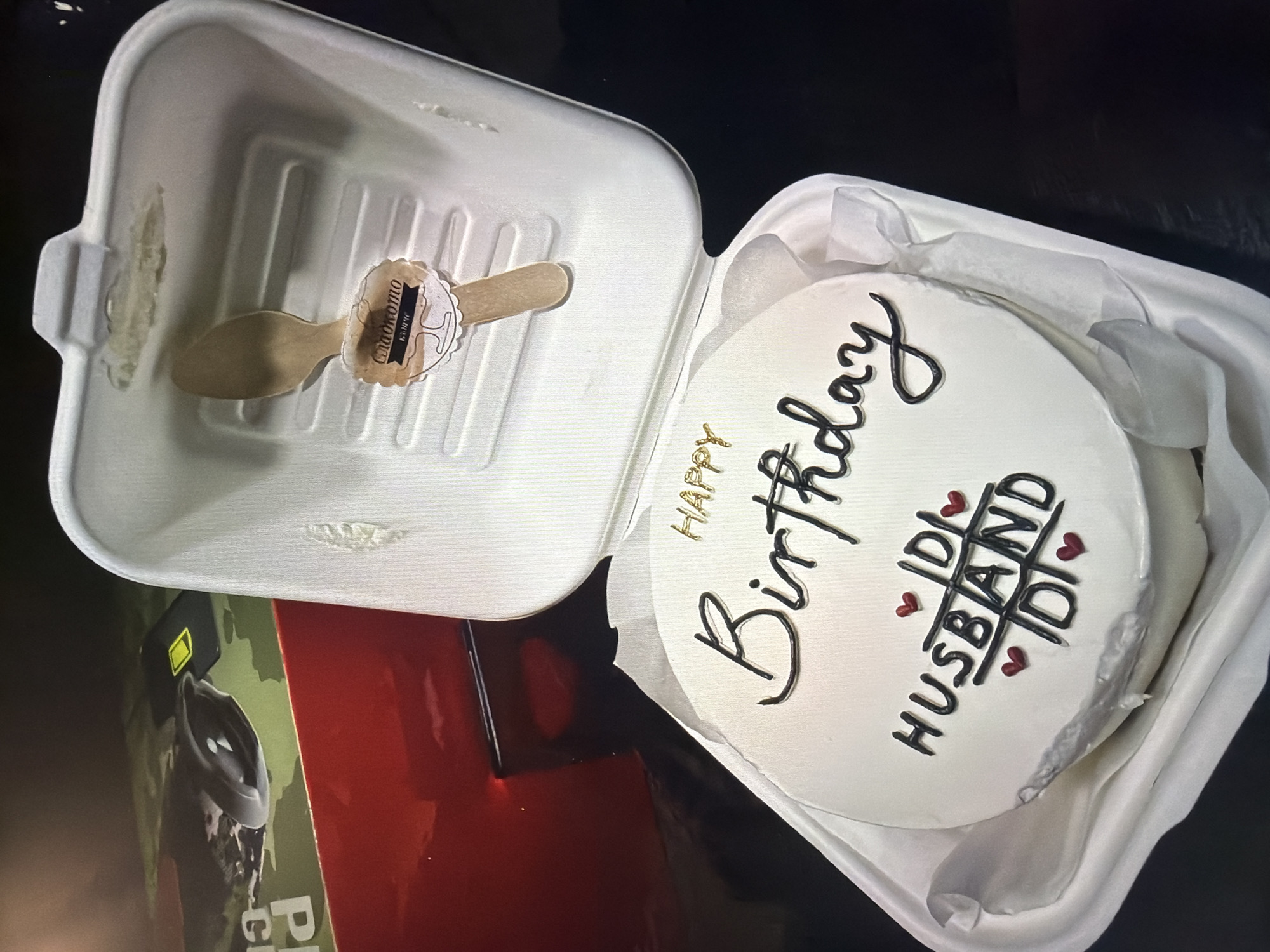 The image shows a white cake with "Happy Birthday" written on it in black and gold icing. Underneath the word "Birthday" are the words "IDI Husband IDI" written in black and separated by red hearts. The cake is in a white takeaway container lined with white tissue paper. Next to the cake container is another open white container with a wooden spoon inside. Part of a red box with a green camouflage pattern is visible in the lower left corner of the image. The exact location in the city of Unknown, Unknown is not discernible from the given information.