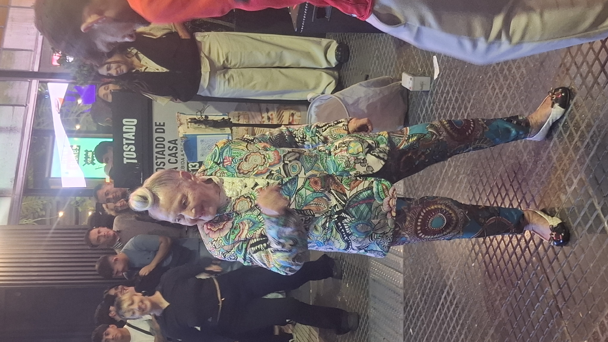 The image depicts a person standing on a textured metal surface, likely a grate. The person is wearing a vividly patterned jacket and pants with intricate designs. Their hair is styled in a bun on top of their head, and they wear a pair of low-cut shoes. A group of people is visible behind them, some appearing to be looking at the person in the foreground, and there is a sign visible on the left with the word "TOSTADO" and some information in Spanish. The setting is an urban outdoor location, likely in Buenos Aires, Argentina, due to the signage in Spanish.