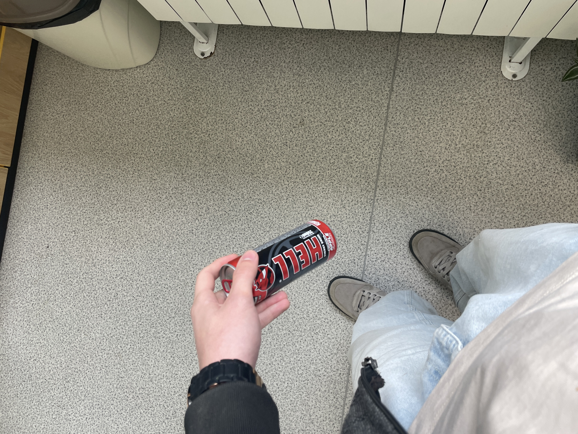 A first-person, overhead shot depicts a person holding a can of Hell energy drink indoors. The person is wearing light blue jeans, grey sneakers, a black jacket sleeve, and a black watch. The background consists of a grey tiled floor, a white radiator, and a portion of a white object (possibly a bin). No information relates to the specific location in Burgas, Bulgaria.