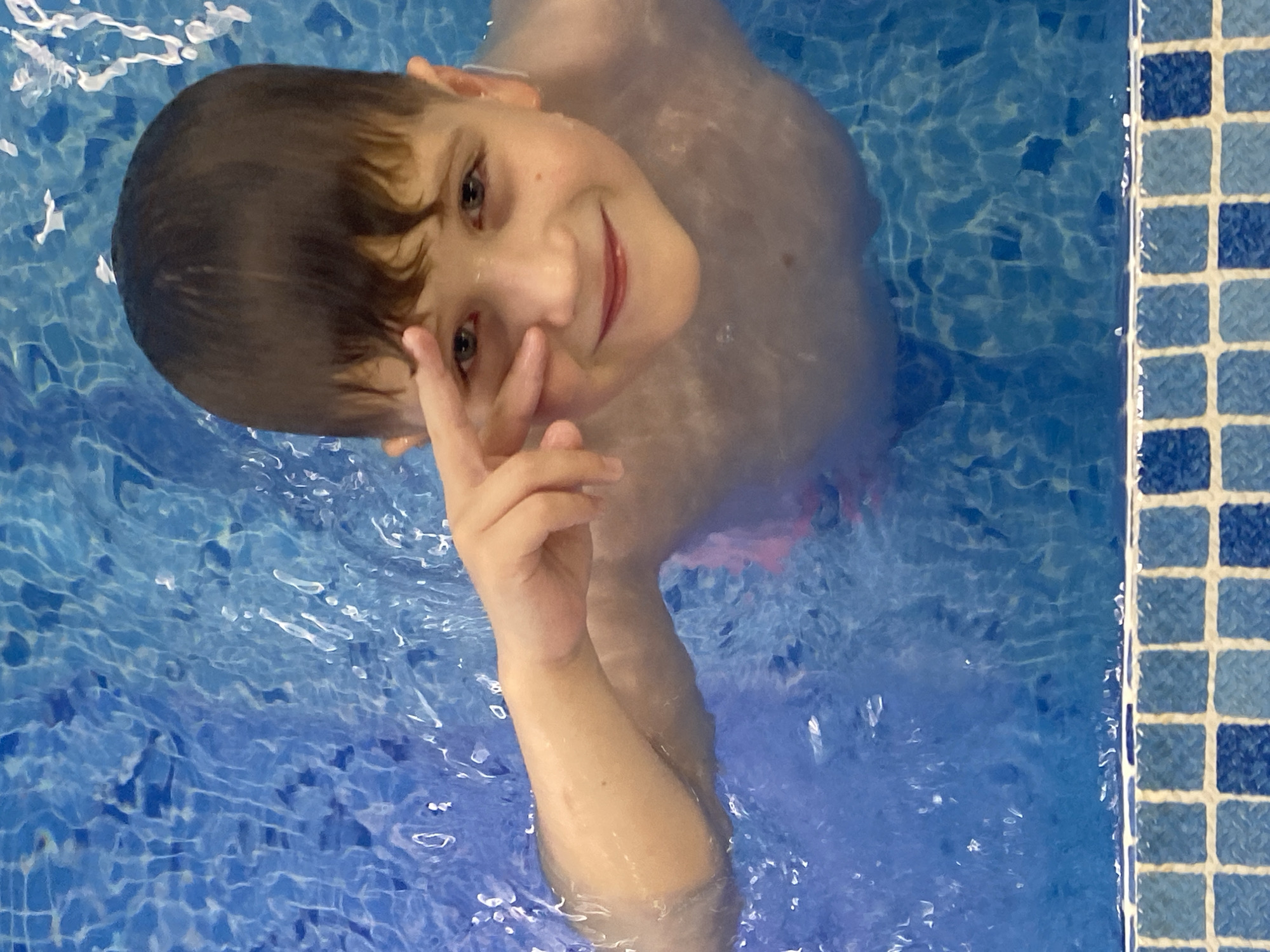 In this medium shot, a young boy with dark hair and grey eyes is submerged in a swimming pool, looking up and smiling at the camera. He is making a peace sign with his right hand, his fingers splayed. His skin is wet, and ripples of water surround him. The pool's surface reflects light, creating a shimmering effect. To the right of the frame, a tiled edge of the pool is visible, with small, square tiles in varying shades of blue. The lighting suggests it could be indoors or late in the day. The boy's expression conveys happiness and playfulness. The scene is set in Votkinsk, Russia. No text is visible in the image.