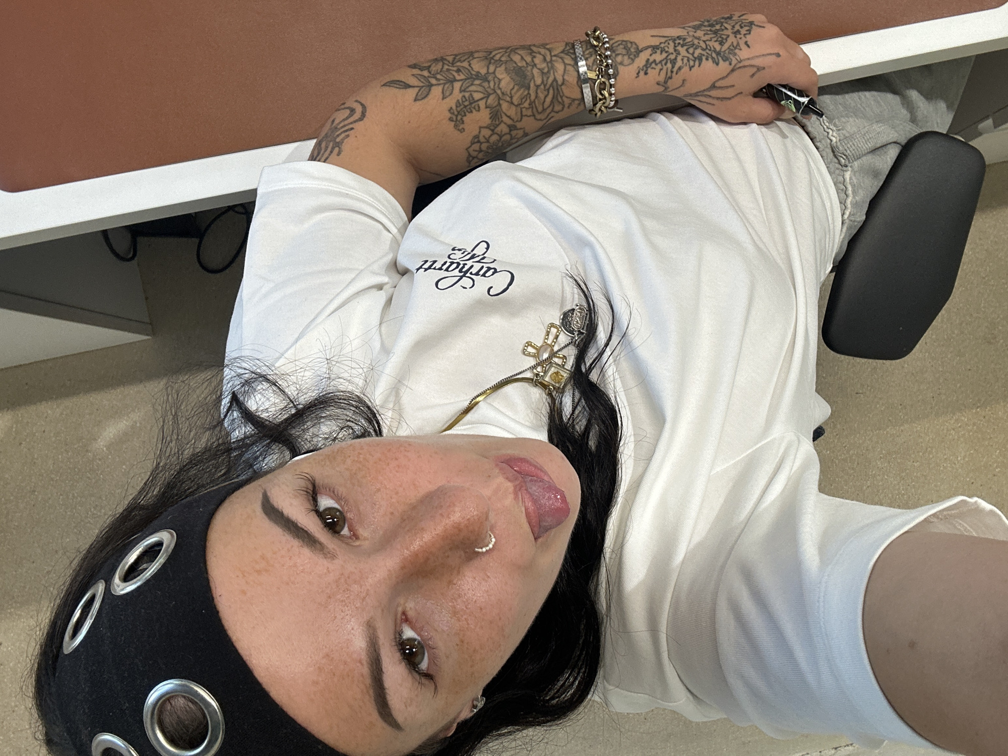A young adult female is seen from a tilted, high-angle perspective. She is sitting at a desk in an indoor environment that could be an office space. The desk is brown and white. She wears a white t-shirt with a Carhartt logo, grey sweatpants, and a black headband with metal eyelets. She also has several bracelets on her left wrist, visible tattoos on her left arm and a gold necklace. She has brown eyes and freckles, a nose ring and is sticking out her tongue. A black pen is in her left hand. Part of a black office chair is visible near her right hip. There is no evidence of the location being in Paris, France.