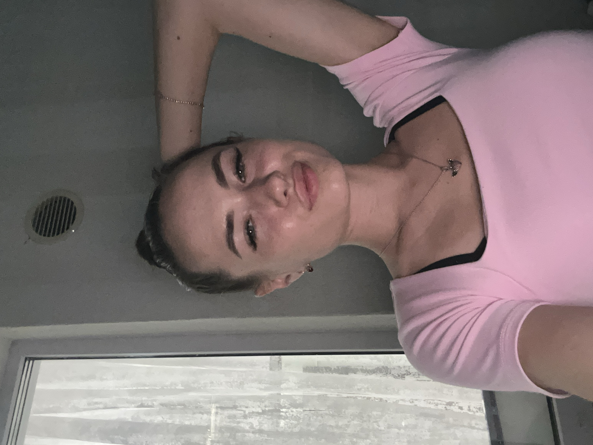 A young woman with her hair in a bun, wearing a pink t-shirt and a necklace, poses for a selfie. She has her left arm raised behind her head, and her right arm is resting on her hip. The background shows a window with sheer curtains to the left and a gray wall with a vent on the right.