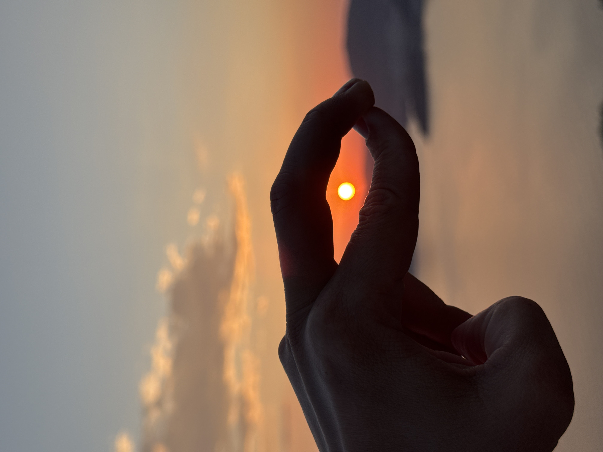A low-angle, close-up shot captures a human hand making a small "O" shape with its fingers, framing the setting sun. The bright yellow sun is centered within the "O," silhouetting the hand against the sky. The sky fades from a bright orange near the horizon to a lighter blue-grey higher up. Soft, blurred cloud formations are visible in the background. The location is Boreti, Montenegro.