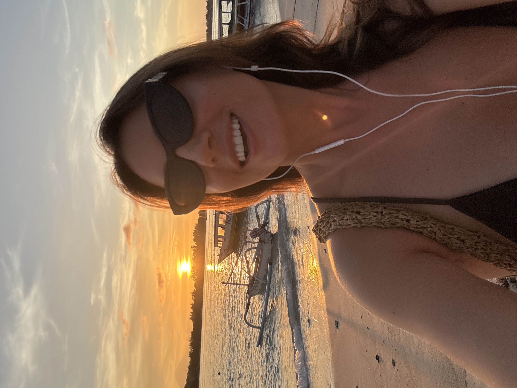 A woman is taking a selfie on a beach during sunset. She is smiling, wearing sunglasses, and has earphones in. The sun is setting over the ocean, with a small outrigger boat visible in the water near the shore. The location is Unknown, Indonesia.