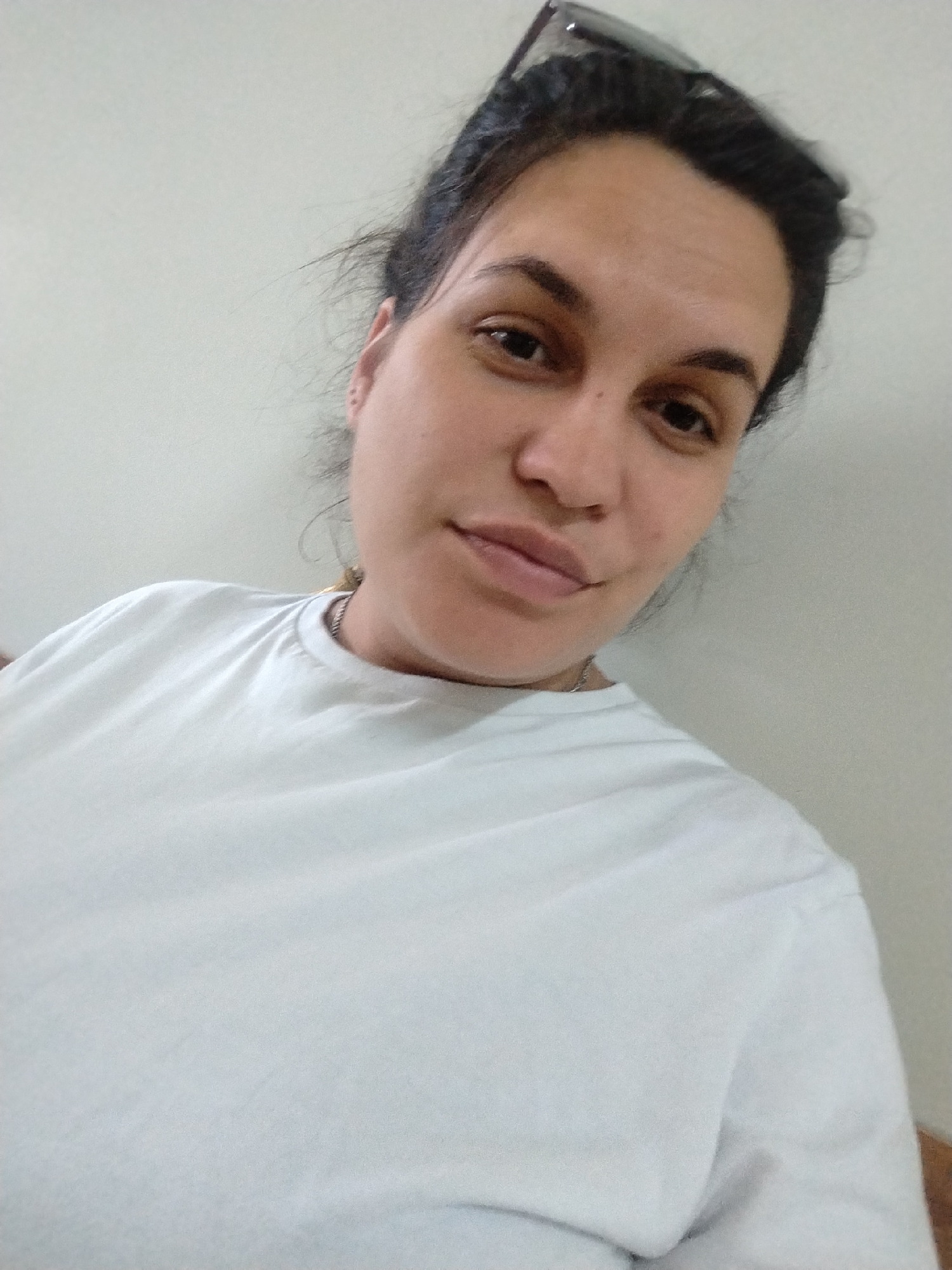 A woman is centered in the image, posing for a selfie. She has dark hair pulled back from her face, light skin, and is wearing a plain white T-shirt. Sunglasses rest on the top of her head. The background is a plain, light-colored wall. There is no discernible information to indicate the location is in Фоки, Russia.