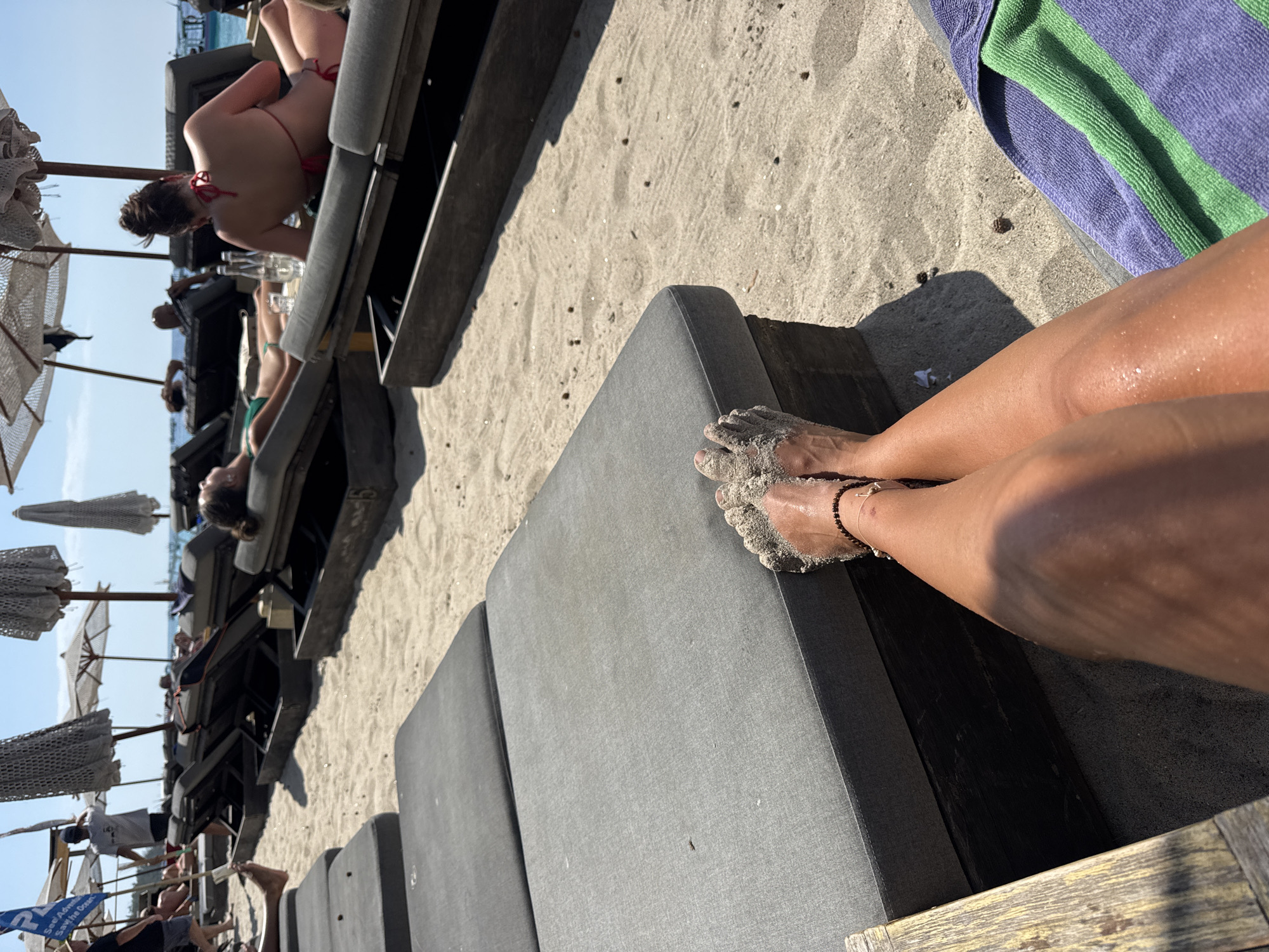 A close-up, POV shot shows a person's legs and feet covered in sand, resting on a gray beach lounge chair. Their feet are adorned with a beaded anklet. Behind them, the sandy beach is lined with more lounge chairs, some occupied by sunbathers. Beach umbrellas provide shade. In the distance, the ocean is visible under a clear blue sky. The overall scene depicts a relaxing day at the beach.