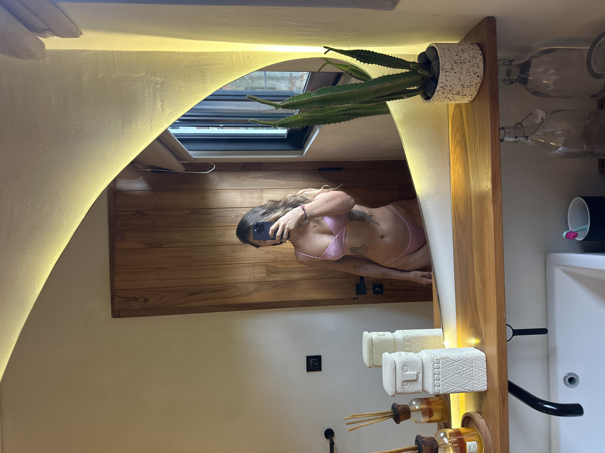 The image, captured in North Kuta, Indonesia, depicts a mirror selfie taken by a woman in a modern bathroom. The woman, positioned centrally, is wearing a light pink two-piece bikini or lingerie set. Her face is obscured by the smartphone she holds to take the photograph, with tattoos visible on her left arm and torso.
The bathroom features light-colored walls and a distinctive arched mirror framed by warm strip lighting. Below the mirror, a wooden vanity or shelf extends across, holding an assortment of items. On the left side of the vanity is a potted cactus plant in a speckled white pot. To the right, two intricately carved, light-colored stone statues resembling figures are displayed. Further along are two reed diffusers with amber liquid and wooden stoppers, and two clear glass bottles with swing-top caps. A white sink is partially visible on the far right. A black electrical outlet or switch is mounted on the wall below the mirror.
The reflection in the mirror reveals a large skylight or window with horizontal blinds, offering a glimpse of what appears to be architectural elements or rooftops outside. The room visible in the reflection also shows wooden plank flooring. The overall scene suggests a stylish and comfortable interior space. No legible text is visible in the image.