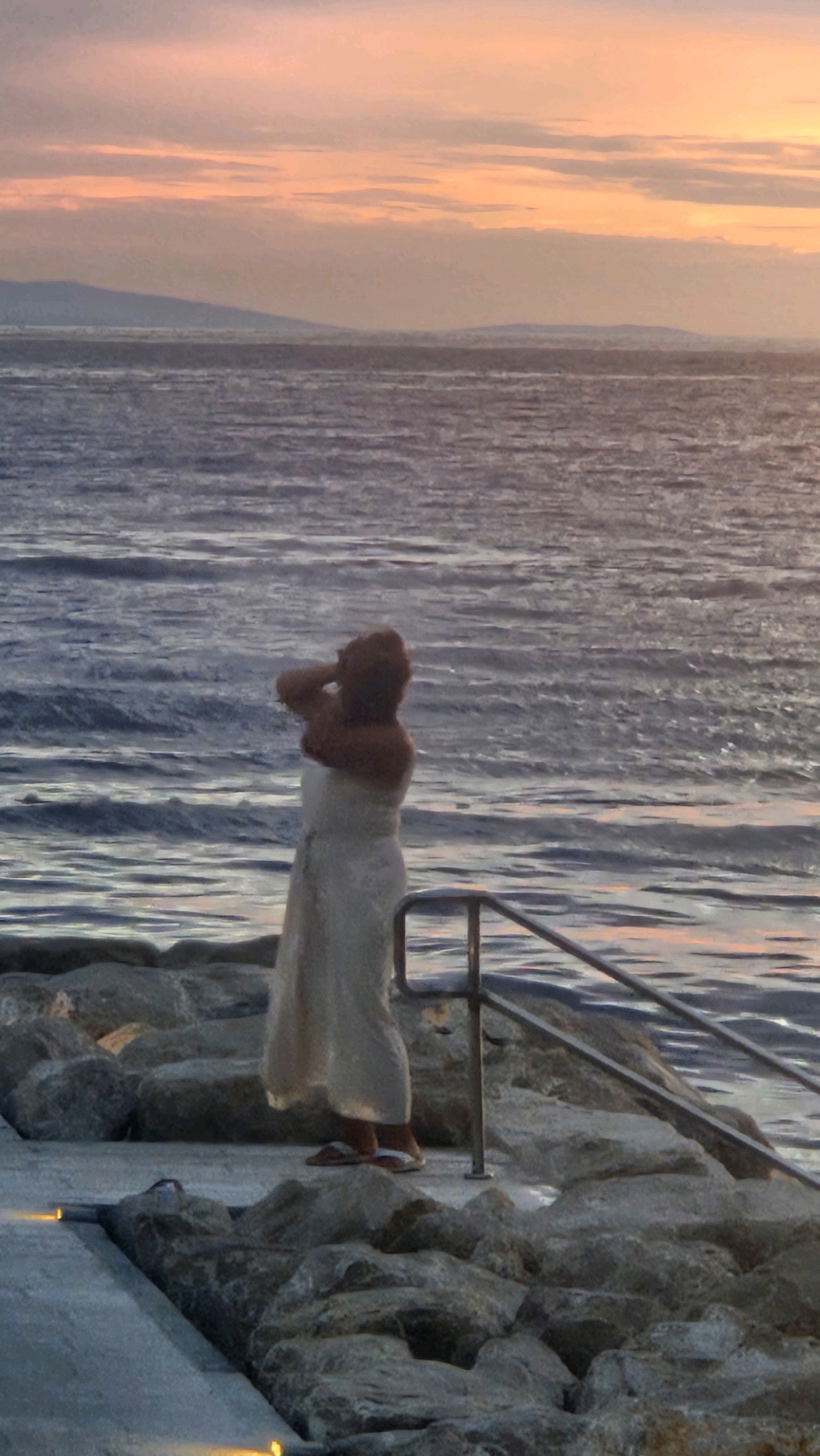 A person stands on a rocky embankment overlooking the sea at sunset. The person, seen from behind, is wearing a light-colored dress and has their hands raised to their head. The sea is choppy with ripples reflecting the pink and orange hues of the sky. In the distance, a hazy coastline is visible. The embankment is made of large, irregular stones, and a metal railing runs along its edge. The scene appears to be in a coastal area.