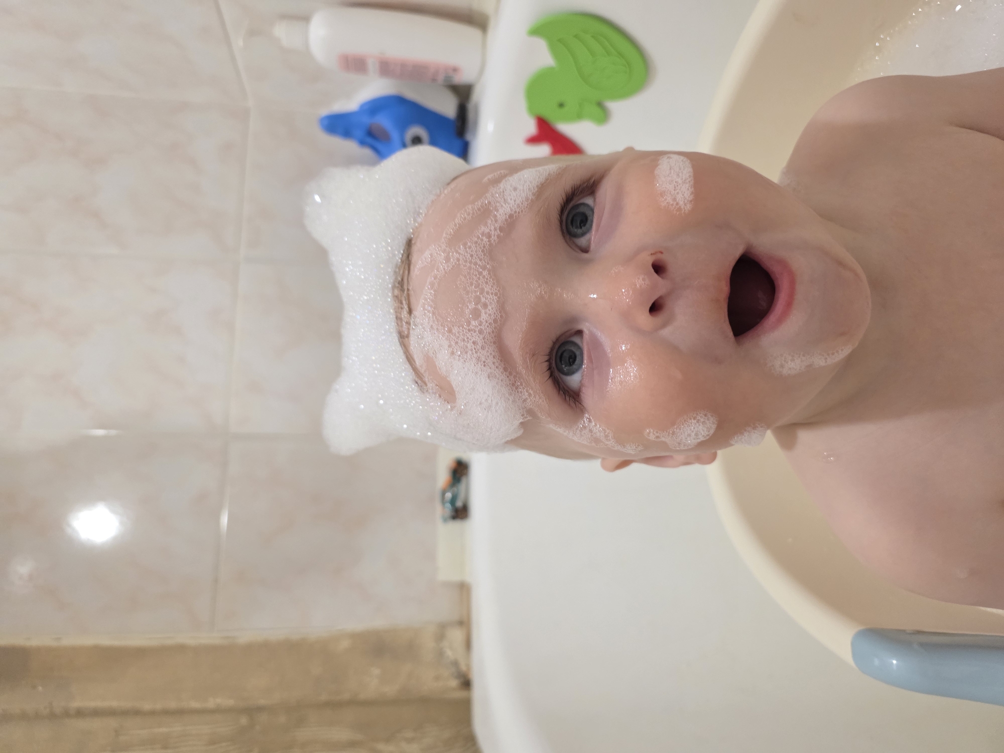 A close-up, rotated image shows a baby in a bathtub, covered in soap bubbles. The baby has blue eyes and an open mouth. In the background, there are bathroom tiles, a bottle of shampoo, and bath toys. The location of the photo within Chaikovsky, Russia cannot be determined from the image.