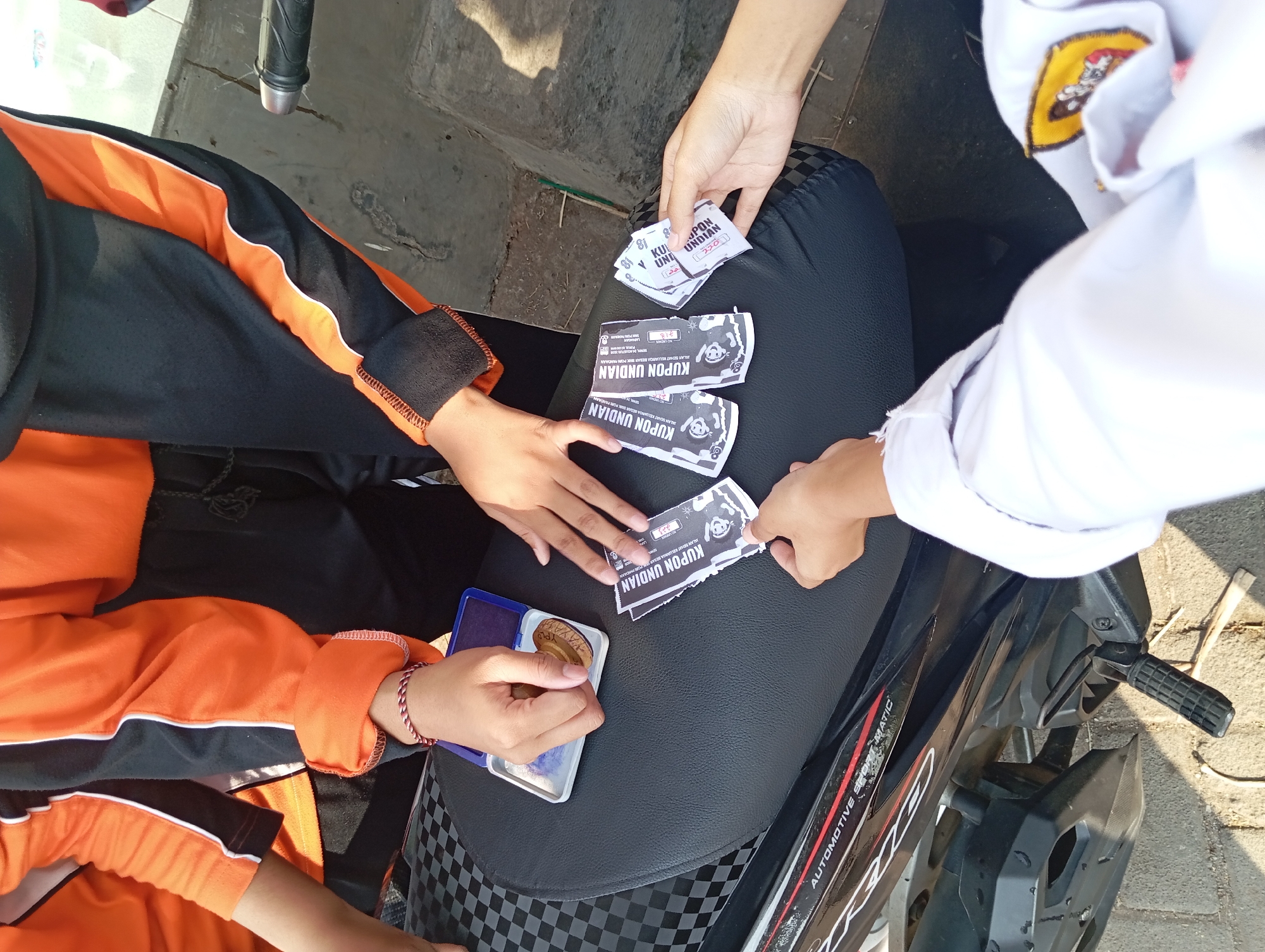 In Pandaan, Indonesia, a group of individuals is gathered around a motorcycle seat, engaged in an activity involving coupons and a stamp. A person in the foreground, wearing a white garment possibly a school uniform, is reaching out to receive coupons. To their left, another individual with an orange and black jacket is holding a stamp pad and a stamp. Scattered on the black motorcycle seat are several coupons, some of which appear to be marked with the stamp. The overall environment suggests an outdoor setting, with a paved ground and sunlight indicating it's daytime. The presence of a motorcycle points towards a public area or a street in the city.