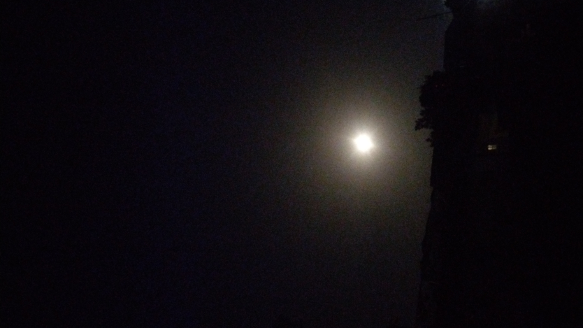 The image depicts a night scene in Jos, Nigeria. A bright light source is visible in the center, possibly a streetlight. The surrounding environment is dark, with vague shapes of trees and a building silhouette visible to the right. No people are visible. The lighting suggests a relatively high exposure setting on the camera, which may obscure details in the darker areas.