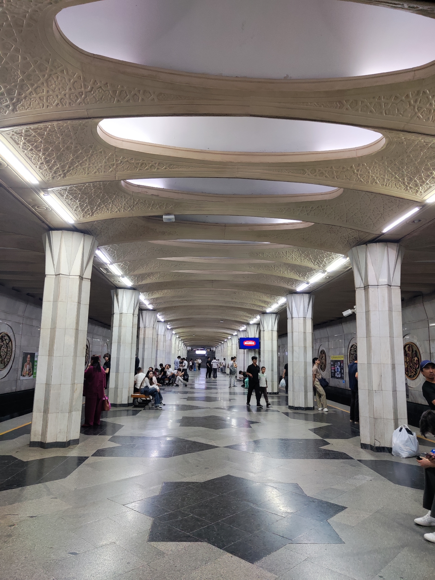The image shows an interior view of a metro station in Tashkent, Uzbekistan. The station features ornate architecture, with a long, arched ceiling adorned with geometric patterns and recessed lighting. A series of large, fluted pillars line the platform, supporting the ceiling. The floor is tiled with a distinctive pattern of dark gray and light gray geometric shapes.
Several people are visible on the platform, appearing to wait for a train or move through the station. Some are seated on benches, while others are standing. The individuals are dressed in casual clothing. On the walls, there are decorative panels with circular motifs and posters. A digital display board with blue lighting is also visible further down the platform. The overall impression is one of a grand and well-maintained public transportation hub.