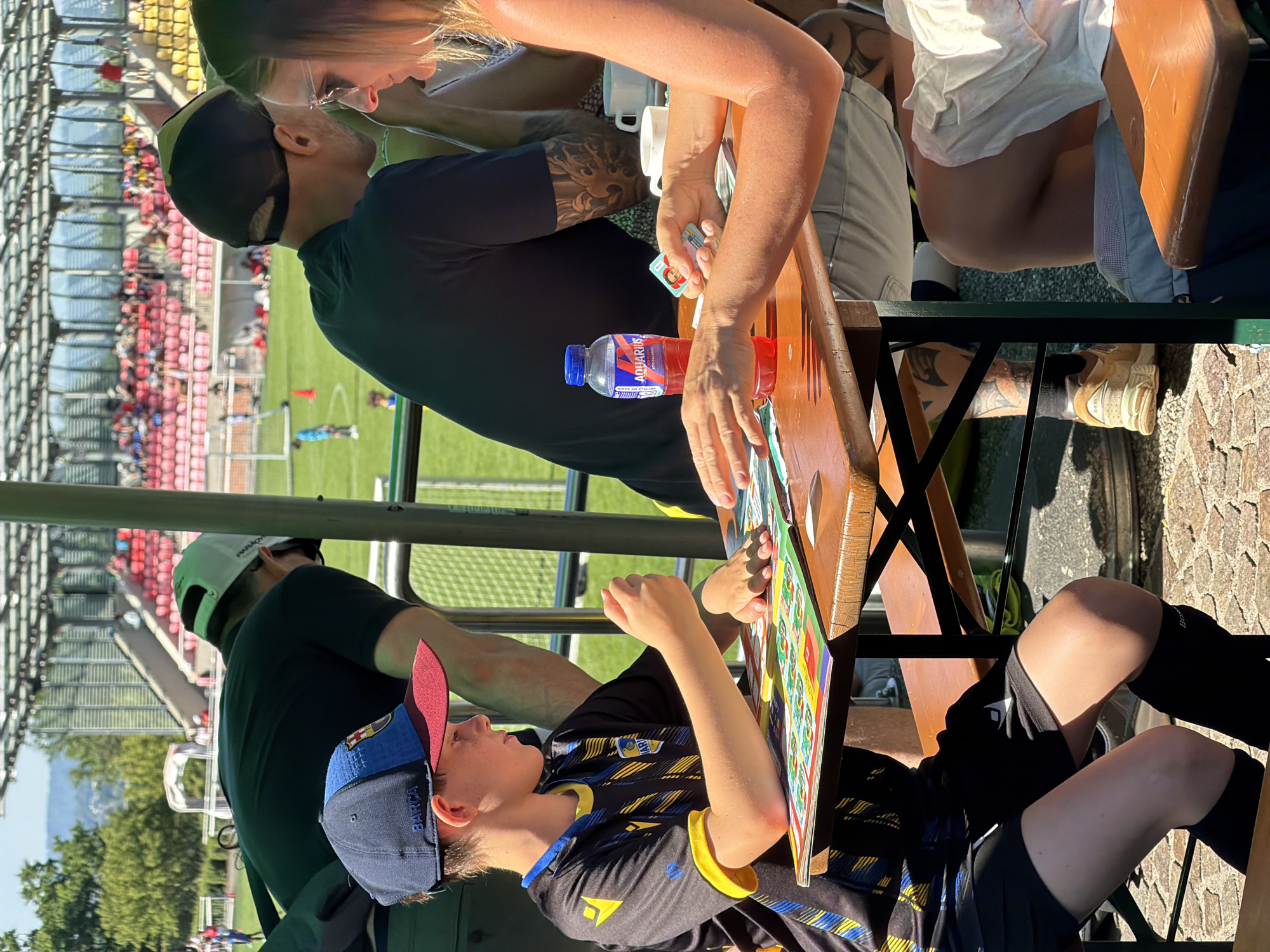 A family is sitting at a table outdoors at a stadium in Fentange, Luxembourg, with a soccer game taking place in the background. A boy wearing a soccer uniform and a Barcelona cap is playing a game on a board. A woman with long blonde hair and a man with a black baseball cap and arm tattoos are sitting across from the boy, playing cards. A water bottle is on the table. A person with a white baseball cap is sitting behind the boy. Red stadium seating and a soccer field are visible in the background.