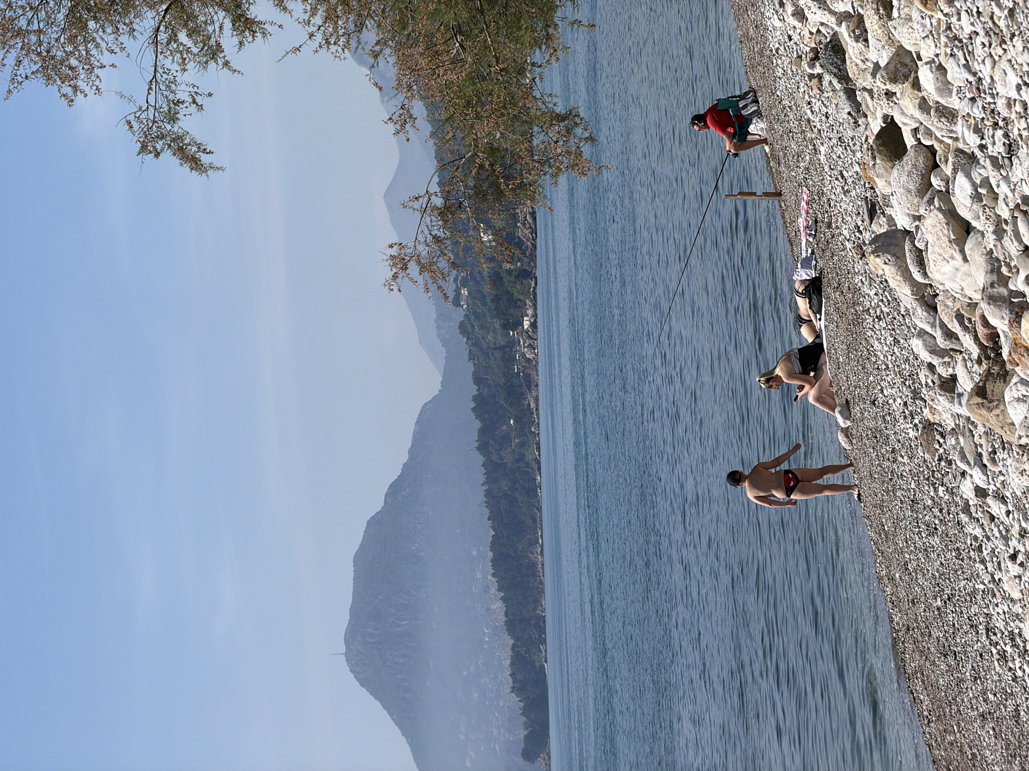 This image captures a serene coastal scene at a pebble beach in Bar, Montenegro, on a clear, sunny day. The image is oriented incorrectly, but when viewed correctly, it shows a wide expanse of calm, blue-green sea meeting a light-colored, rocky and pebbly beach. In the background, layers of hazy blue mountains rise, topped by a light blue sky.
Several people are enjoying the beach. On the right side, a man wearing a red t-shirt, dark shorts, and sunglasses is actively fishing with a long fishing rod, which rests on a wooden stand. He is standing near the water's edge. Further along the beach to his left, another person is lying down, partially visible, wearing dark swimwear. Slightly closer to the foreground and to the left of the fisher, a person with light hair, possibly blonde, is seen sitting or lying on the pebbles, looking down at what appears to be a mobile phone. Closest to the foreground, a man in black and red swim trunks stands facing the water, his back partially visible, near the gentle lapping waves.
Overhanging the top left portion of the frame are branches of a leafy tree, providing a natural frame to the scene. The overall atmosphere is one of relaxation and leisure, typical of a summer day at the beach. No discernible text is visible on any objects or clothing.