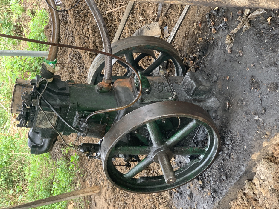 The image shows a large, green, industrial engine, lying on its side, partially obscured by green foliage in the foreground. The engine has a large, spoked, black wheel. The engine is lying on a bed of dark, wet, muddy soil. The engine appears to be in disrepair, with parts missing or damaged. The image was taken at coordinates (latitude: 7.119, longitude: 5.362) in the city of Egbeda, Nigeria.