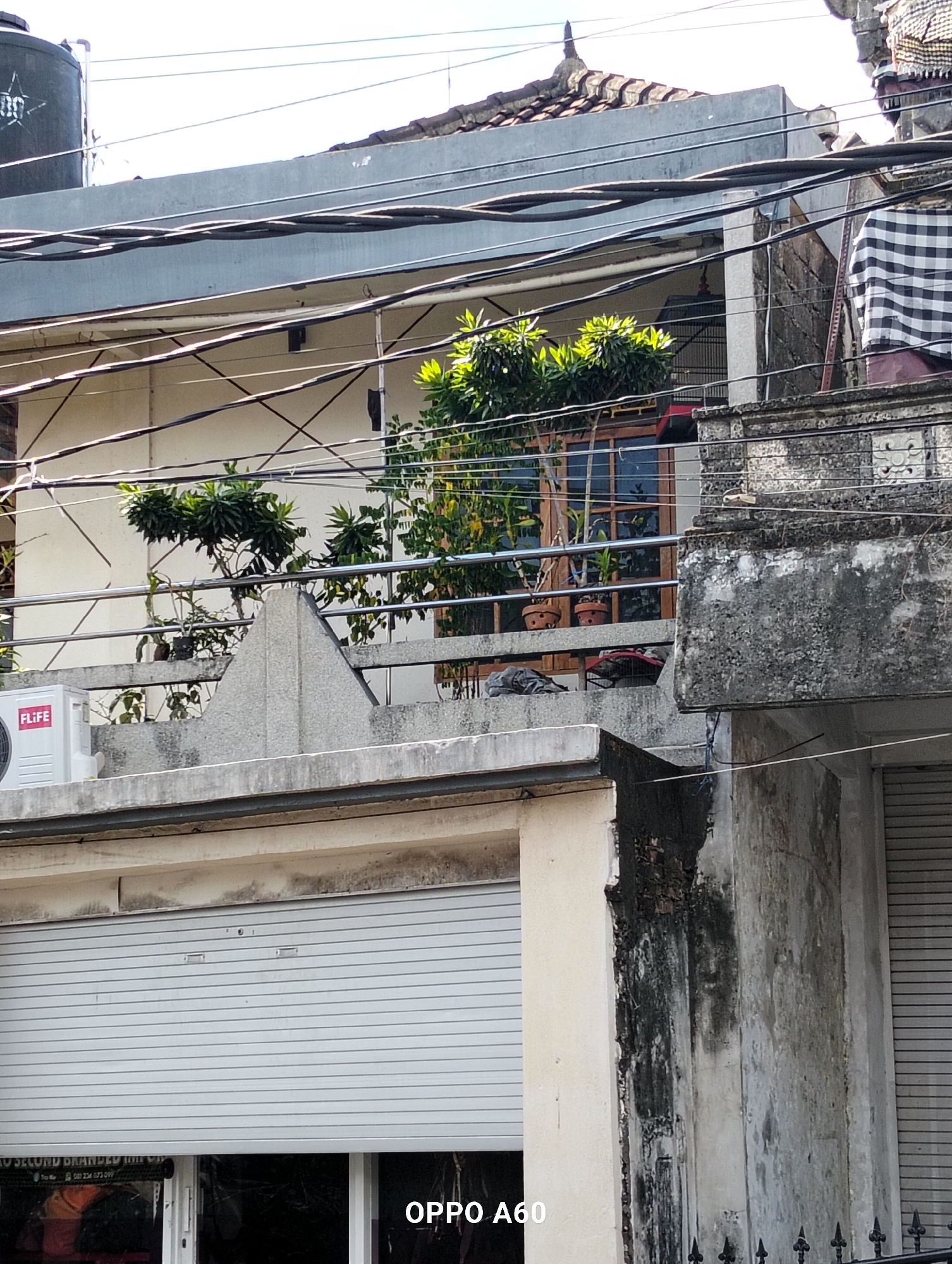 The image shows a close-up view of an urban building exterior in Denpasar, Indonesia, likely on a sunny day. A prominent feature is a dense network of overhead electrical wires crisscrossing the frame. Below these wires, on a balcony or terrace, several potted plants are visible, including some with bright green foliage. An air conditioning unit with the "FLiFE" logo is mounted on a concrete ledge. The building's facade is textured and shows signs of weathering and some staining. A rolled-down metal shutter is visible on the lower part of the building, with a sign partially obscured at the bottom left. A traditional Balinese decorative element, possibly a carved stone, is visible on an upper section of the building to the right, draped with a black and white checkered cloth. The overall scene depicts a cluttered but typical urban environment with a mix of modern amenities and traditional architectural details.