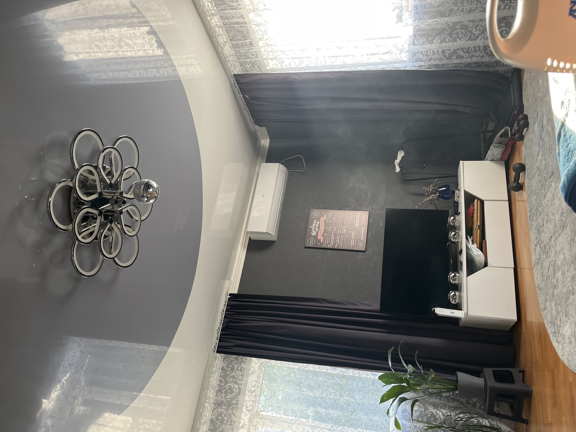 This image depicts the interior of a living room, likely in an apartment. The room features a modern, multi-level ceiling with a glossy gray upper section and white lower sections, creating a dynamic effect. A distinctive contemporary chandelier with multiple circular elements and a disco ball hangs from the ceiling. The walls are painted a dark, textured gray.
Dominating the wall is a large, flat-screen television mounted above a white media console. On the console are several decorative silver spherical objects. To the left of the television, a large potted plant with green leaves is placed on a small black stool. To the right of the media console, a treadmill is partially visible behind dark gray curtains, suggesting a home gym setup.
The room is illuminated by natural light filtering through lace curtains on the windows, casting patterned light across the floor and ceiling. A white air conditioning unit is mounted high on the wall above the television. The floor appears to be made of polished wood, and a patterned gray rug is visible in the foreground.
The presence of a treadmill and dumbbells indicates that physical fitness is a part of the occupants' lifestyle. The overall aesthetic is modern and functional, with a focus on a clean and somewhat minimalist design, punctuated by the statement chandelier and the dark gray walls.
Given the information provided, this scene is located in Saratov, Russia.