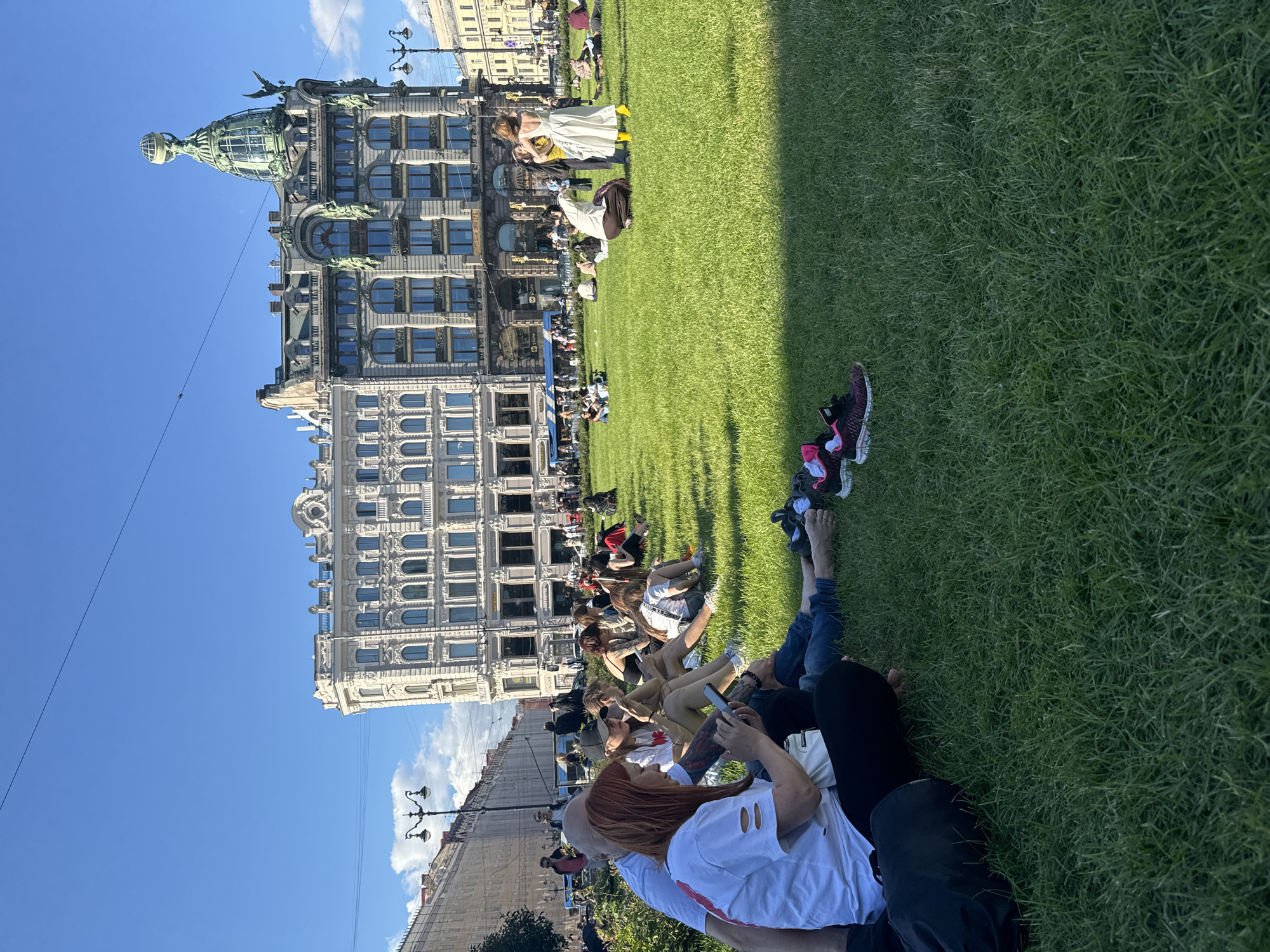 In Saint Petersburg, Russia, on a sunny day with a clear blue sky, a group of people are sitting on a well-maintained green lawn. The lawn is in front of several grand, historic buildings. The most prominent building, located in the background and center, is the Singer House, characterized by its unique dome topped with an eagle and globe. To the left and right of Singer House are other ornate buildings with classical architectural features, including detailed facades, arched windows, and decorative cornices.
In the foreground, a group of people are seated on the grass, some facing away from the camera, while others are partially visible from the side or back. A woman with red hair, wearing a white t-shirt, is looking at her phone. Next to her, a person with a black cap is also engaged with their phone. Further back, more individuals are scattered across the lawn, some in pairs or small groups, with a few standing. Scattered on the grass in the foreground are several pairs of athletic shoes. In the mid-ground, closer to the buildings, a few people are standing and interacting. The scene appears to be a public gathering or leisure activity in a city square or park.