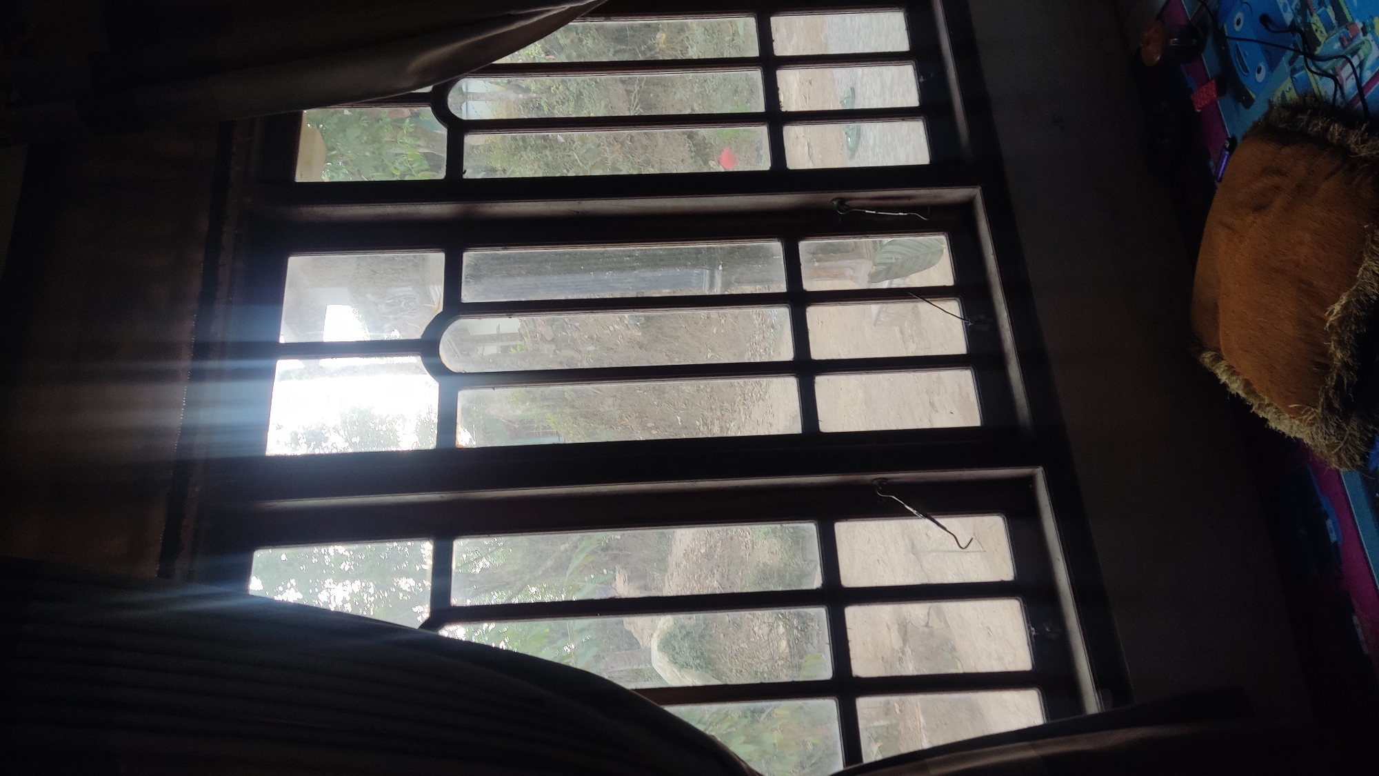 The media file shows a view through a large, three-pane window. The window is framed with dark wood and has horizontal and vertical dividers creating multiple rectangular panes. Sunlight streams through the left side of the window, creating visible light rays that illuminate the interior space. Outside the window, a somewhat overgrown and dusty environment is visible, with some greenery and soil.
The interior appears to be a dimly lit room. On the right side of the frame, a corner of a bed or couch is visible, covered with a colorful patterned fabric, and a brown, furry cushion. A thin, dark curtain partially covers the left side of the window.
The overall impression is one of a domestic interior looking out onto an outdoor scene, with the sun's rays indicating it is likely daytime, possibly morning or afternoon, depending on the direction the window faces. The scene evokes a sense of quiet observation, with the sunlight adding a touch of warmth and perhaps nostalgia to the otherwise dim setting. There is no visible text in the media file. The location context suggests this scene is in Ngadiharjo, Indonesia.