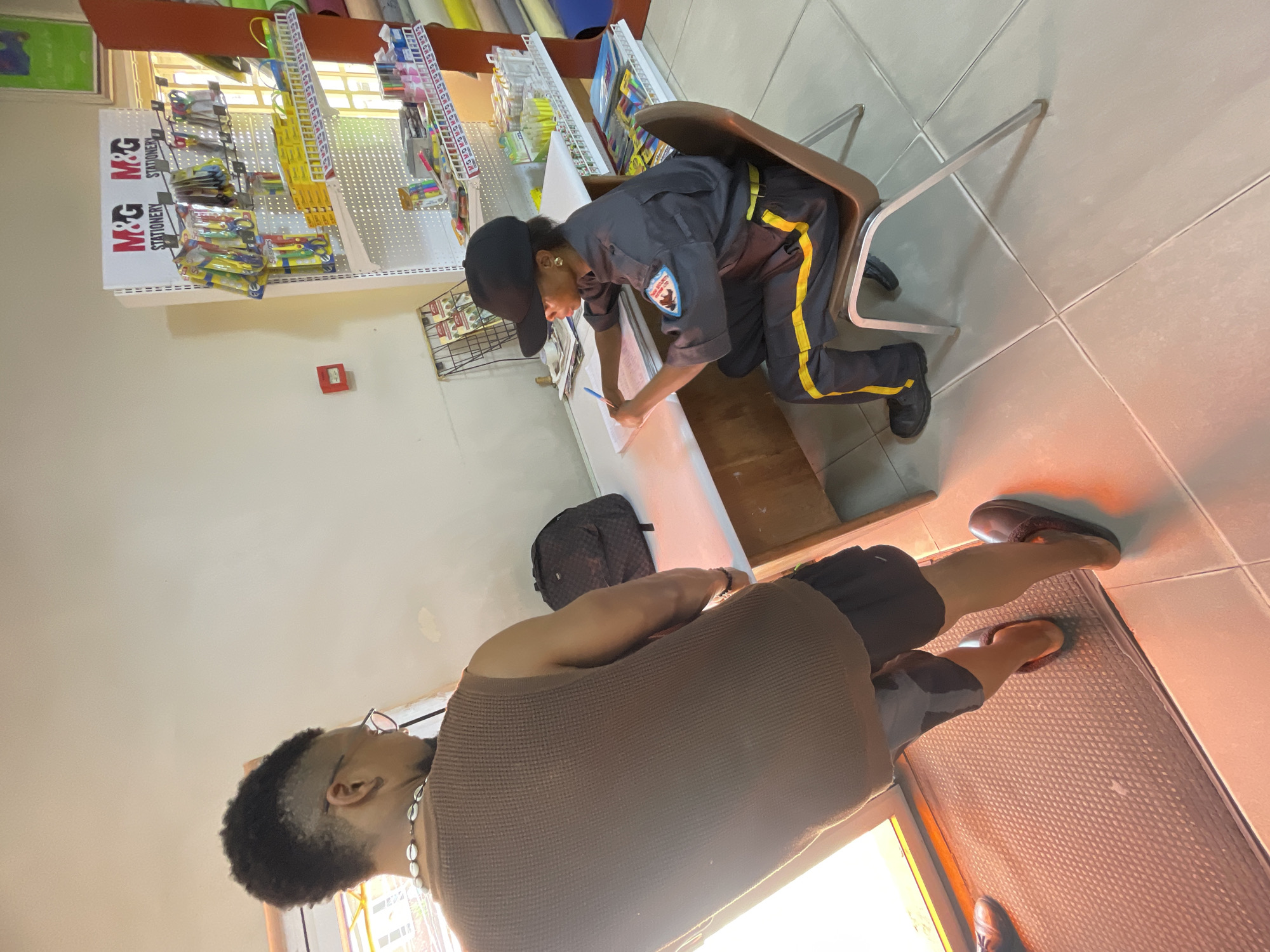 This indoor image depicts a stationery shop or office setting with two individuals. In the foreground, a person with short, dark, curly hair and glasses, wearing a brown waffle-knit top and a shell necklace, stands on a textured mat, facing generally towards the midground. They are wearing dark slippers.
In the midground, a second person, dressed in a dark uniform with yellow stripes on the trousers and a patch on the left sleeve, and wearing a dark baseball cap, is seated on a brown chair. This individual is actively engaged in writing on a white surface with a pen, suggesting an administrative task, record-keeping, or a transaction.
The background features white pegboard shelves stocked with various stationery items, including pens, scissors, and rulers. A prominent white sign displaying "M&G STATIONERY" is visible. A red fire alarm box is mounted on the wall. A dark backpack is placed on a surface near the foreground person.
The floor is tiled, and the scene is illuminated by what appears to be natural light from a window, casting a warm, slightly reddish hue on the floor in some areas. The setting suggests a functional retail or service environment.