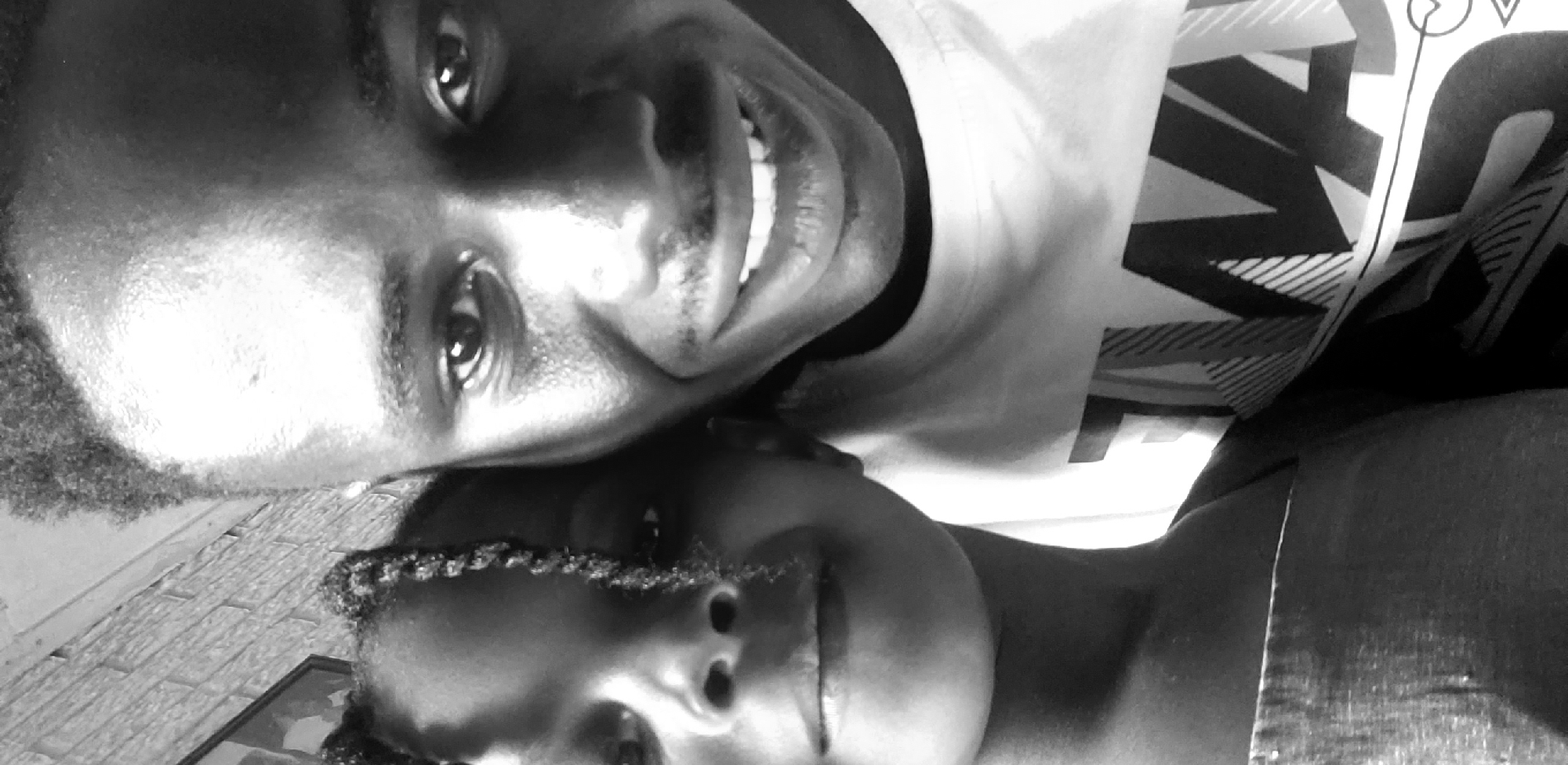 This grayscale image depicts two individuals, an adult male and a child, positioned closely together in a selfie-style shot, located in Narayi, Nigeria. The adult male, on the right side of the frame, has short, dark, curly hair and a broad, open-mouthed smile revealing teeth. He possesses light facial hair, including a mustache and chin beard. He is wearing a light-colored, crew-neck t-shirt adorned with a prominent dark graphic design featuring geometric patterns and large capital letters, including 'T', 'W', and 'A'. The child, positioned on the left, has braided hair, with some braids falling across her face. She exhibits a subtle smile, with her left cheek, nose, and mouth visible. She wears a light-colored garment. Both individuals maintain direct eye contact with the camera. In the blurred background on the upper left, a textured wall, possibly brickwork, and a dark rectangular object, possibly a framed picture, are partially visible. The lighting is ambient, highlighting facial features.