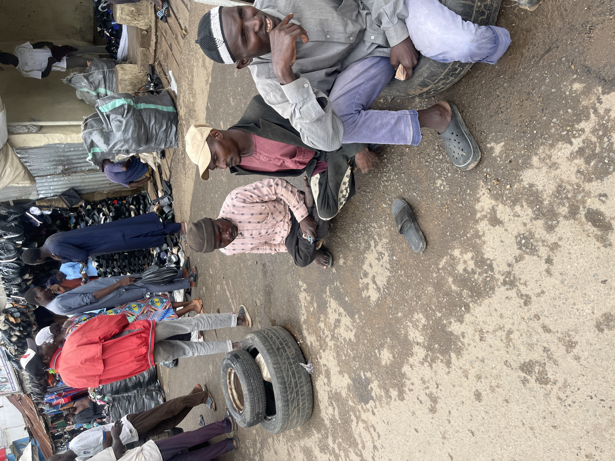 A group of men are gathered in an outdoor market area in Jos, Nigeria. The scene is cluttered with various goods, including stacks of shoes and large bags of what appear to be textiles or other merchandise. In the foreground, three men are seated on the ground. One man, wearing a light-colored button-down shirt and dark pants, is looking towards the right. Another man to his left, dressed in a pink patterned shirt, is looking down. The third man, on the far right, wears a grey shirt and light purple pants and is holding some cash in his hand, looking towards the viewer. Two tires are stacked on the ground to the left of the seated men. The ground surface is a mix of dirt and concrete, with some wet patches. In the background, more market stalls and people are visible, including a man in a red jacket. The overall atmosphere suggests a busy marketplace.