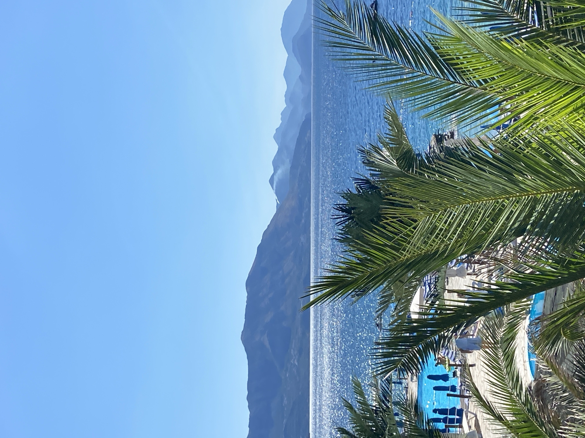 This image depicts a clear, sunny day at a coastal resort, likely in or near Prijevor, Montenegro, given the mountainous landscape. In the foreground, lush green palm fronds partially obscure the view, suggesting a tropical or Mediterranean setting. Beyond the foliage, a vibrant blue swimming pool is visible, surrounded by lounge chairs and umbrellas, indicating a recreational area. People can be seen near the pool and on the beach, engaged in various activities, though their specific actions are not clearly discernible. The middle ground features the sparkling blue sea, its surface rippling under the bright sunlight. In the background, a range of majestic mountains rises against a clear, light blue sky, their peaks fading into a hazy, atmospheric perspective. The overall scene conveys a sense of a serene and inviting vacation destination.