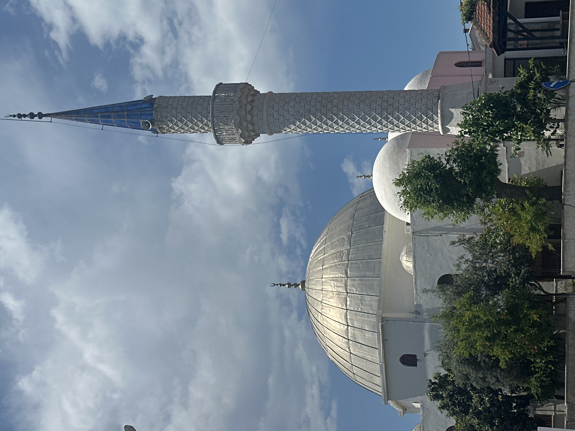 A low-angle, landscape-oriented daytime shot depicts a mosque, identified as the Fethiye Merkez Pasajı Camii mosque located in Fethiye, Turkey. The mosque features a large, silver-colored dome and a slender minaret with a blue conical spire. The minaret is decorated with a textured pattern. The sky is predominantly blue with scattered white clouds. Trees partially obscure the base of the mosque and adjacent buildings. A street sign is visible to the right, among some trees and the building.