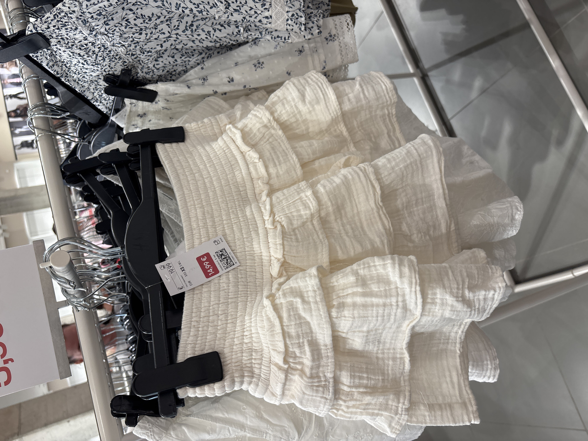 The image shows a close-up of a white ruffled skirt hanging on a clothing rack in a retail store. A price tag is attached to the skirt, displaying "14.99 €" and "19.99 €" along with size information "EUR XXS XS S M L". The background is blurred but suggests other clothing items and store fixtures. The lighting is bright, typical of a retail environment. There is no visible information in the image that places the scene in Orihuela, Spain.