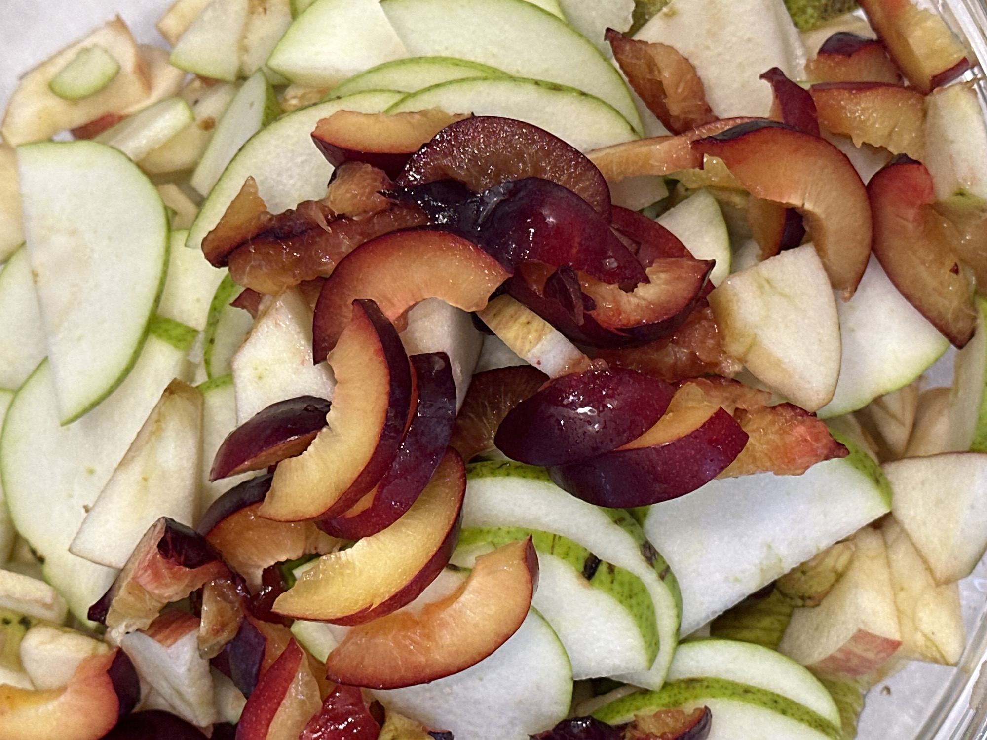 The image is a close-up, overhead shot of a bowl filled with sliced fruits. The fruits appear to be a mix of apples and plums, all cut into various shapes and sizes, some thinly sliced, others in wedges. The apple slices are predominantly white with some showing a hint of green skin, while the plum slices display vibrant shades of deep purple and orange-red flesh. The fruits are jumbled together, creating a colorful and textured composition. There is no discernible environment beyond the contents of the bowl, and no people or actions are depicted. The image provides no information about the city of Nevinnomyssk, Russia, or any other location.