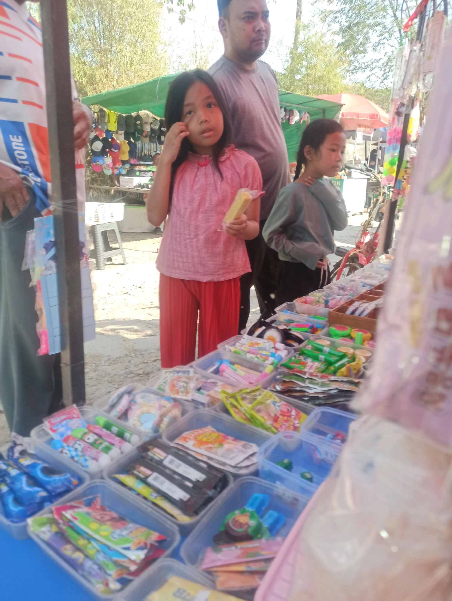 The image depicts an outdoor market scene, possibly in Telagamurni, Indonesia, with a focus on a table laden with various food items and snacks. In the foreground, a young girl in a pink shirt and orange pants stands attentively, holding a yellow snack. To her right, another girl with braided hair, wearing a gray shirt, stands with her hand near her chin, looking to the side. Behind them, a man wearing a gray t-shirt stands observing the scene.
The table is filled with an assortment of packaged goods, including what appear to be candies, chips, and possibly small toys or novelty items. These are arranged in clear plastic containers and on paper packaging. In the background, stalls with green awnings display merchandise, including hats and other items. Sunlight casts shadows on the ground, indicating a bright day. The overall atmosphere suggests a bustling local market environment.