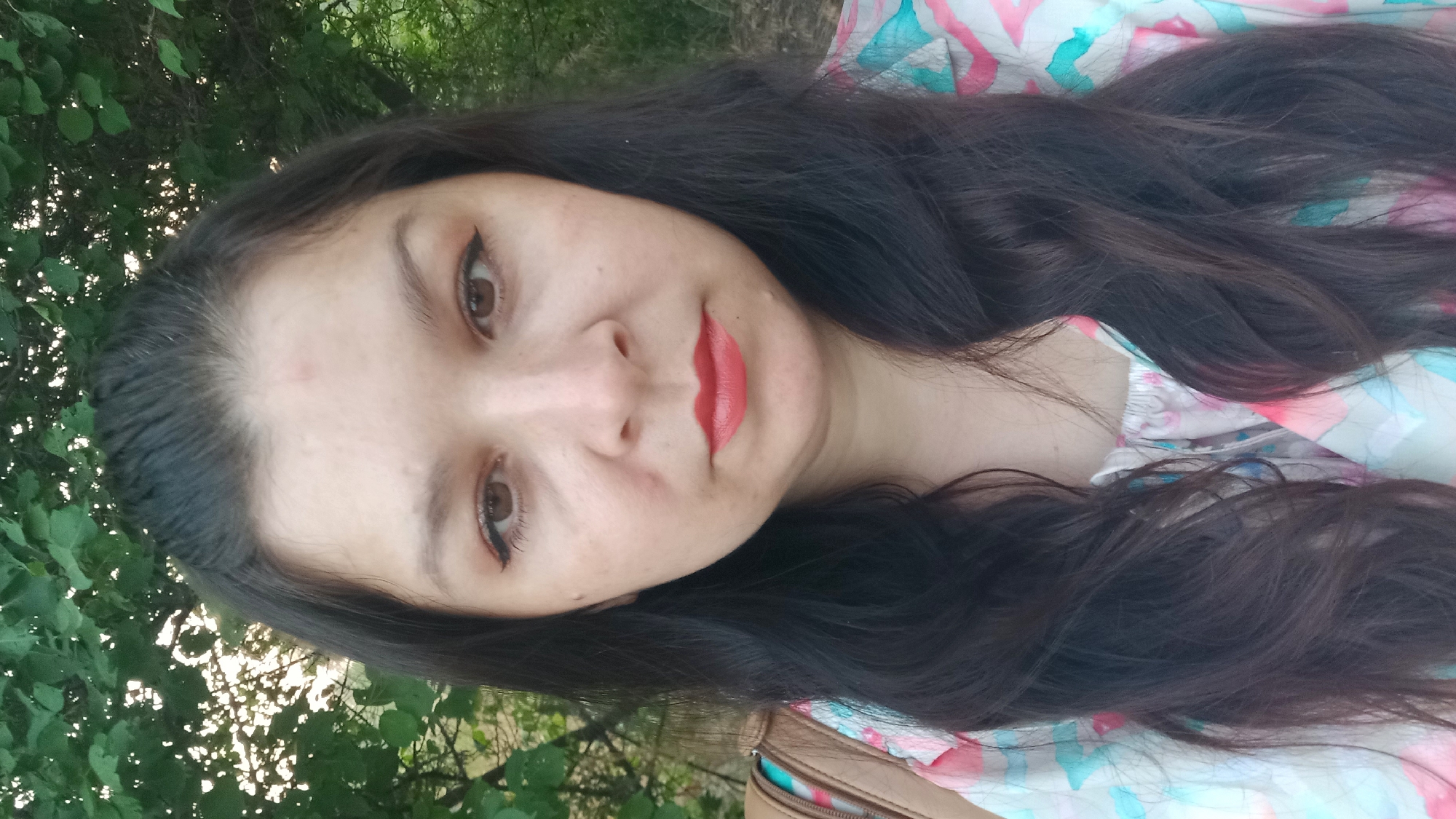 A close-up, waist-up selfie of a woman with dark hair and brown eyes is shown. She is outdoors, with greenery and trees in the background, suggesting a park or natural setting. She is wearing a light-colored shirt with a watercolor-like pattern of pink, blue, and white. Her lips are colored with a coral-red lipstick. The lighting suggests it is daytime. The scene is set in Neopalymivka, Ukraine.